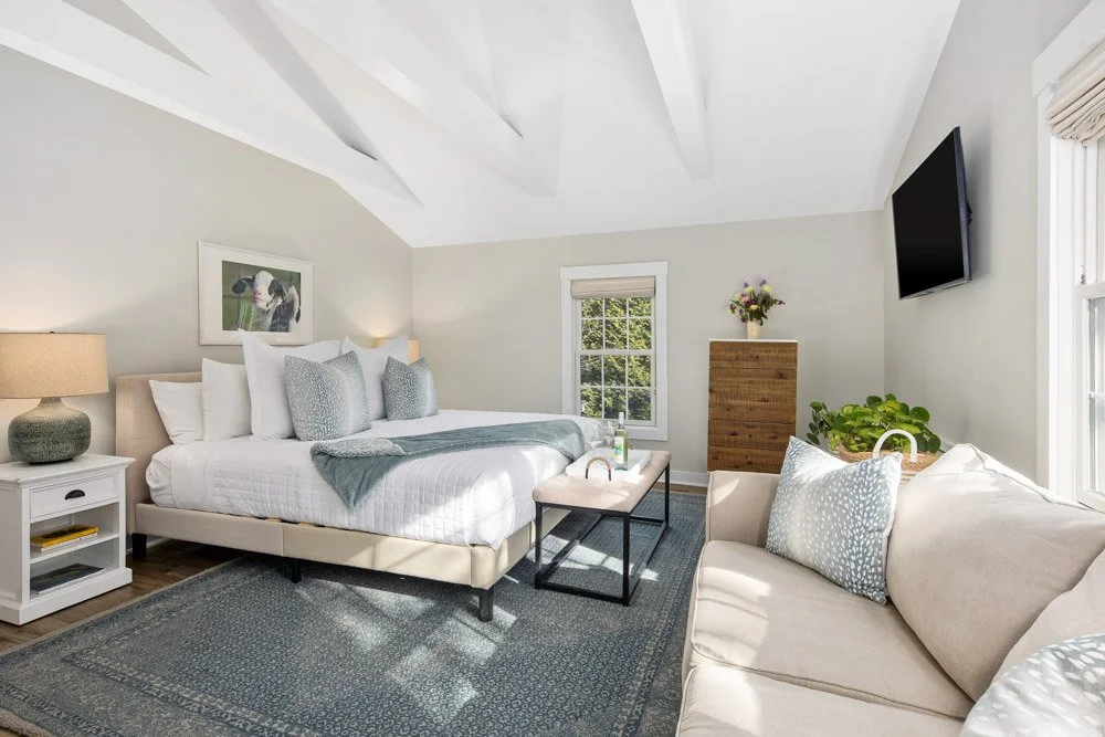 Bright bedroom with a sloped ceiling, white walls, a large bed with white linens and decorative pillows, a beige sofa with pillows, a window with blinds, a dresser with flowers, a wall-mounted TV, and a rug on wooden floor.