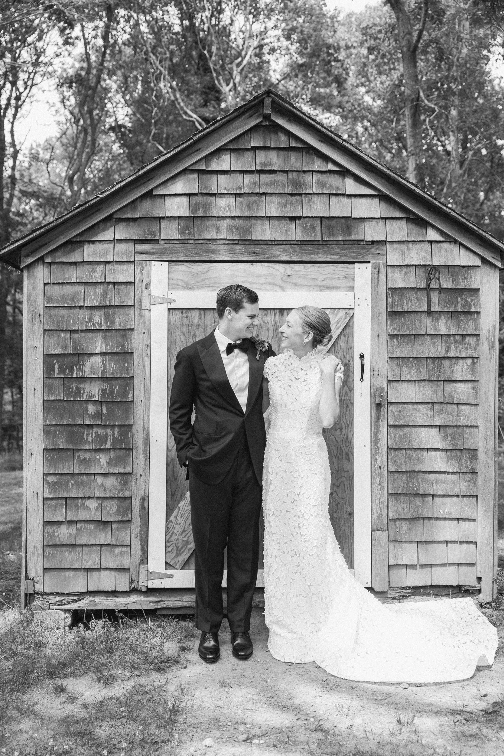 A black and white photo of a bride and groom standing in front of a wooden shed during a wedding at Lambert's Cove Inn on Martha's Vineyard. The groom is wearing a tuxedo and the bride is in a wedding gown, both smiling and looking at each other.