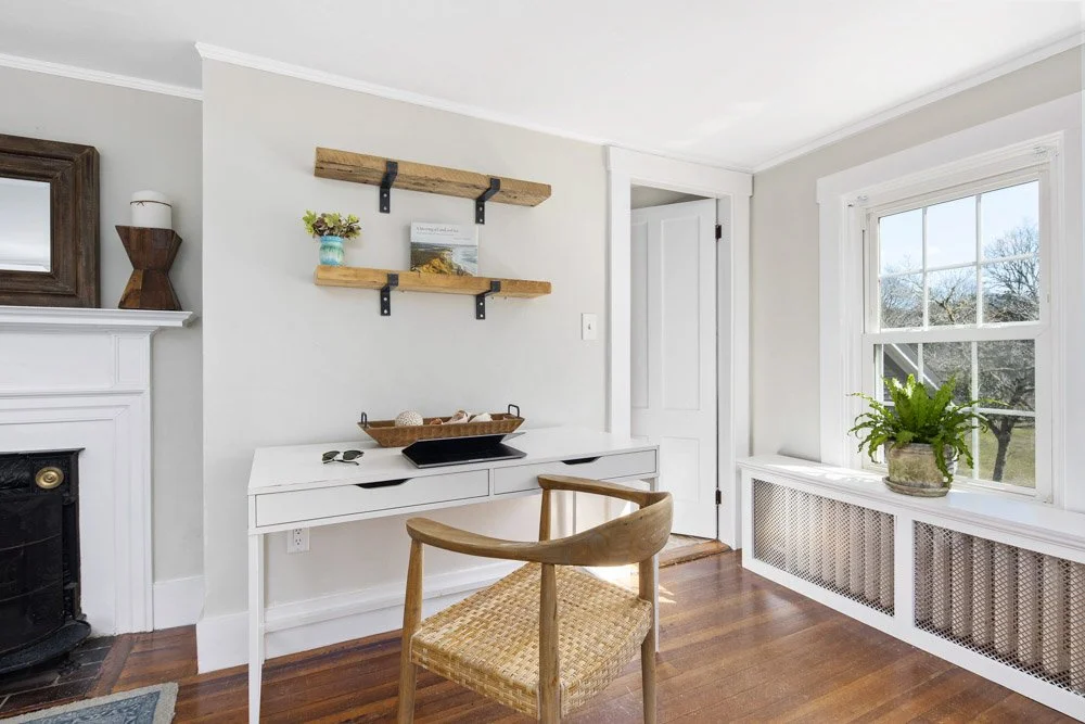 Living room at Lambert's Cove Inn with white walls, a window with a view of trees, a white desk with a wicker and wood chair, two wooden shelves with decorative items, and a fireplace with a mirror and a wooden vase on the mantle.