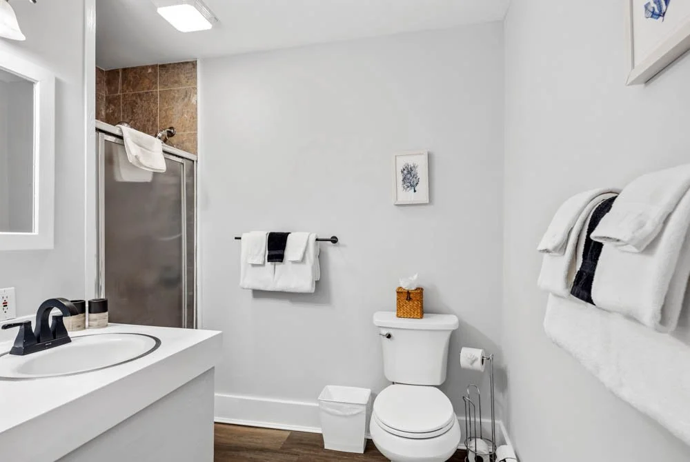 A clean, modern bathroom with a white vanity, a toilet, and a glass shower with brown tile accents. White towels are neatly hung on a towel rack and hooks, with a framed picture on the wall. There is a small trash can and tissue box near the toilet.
