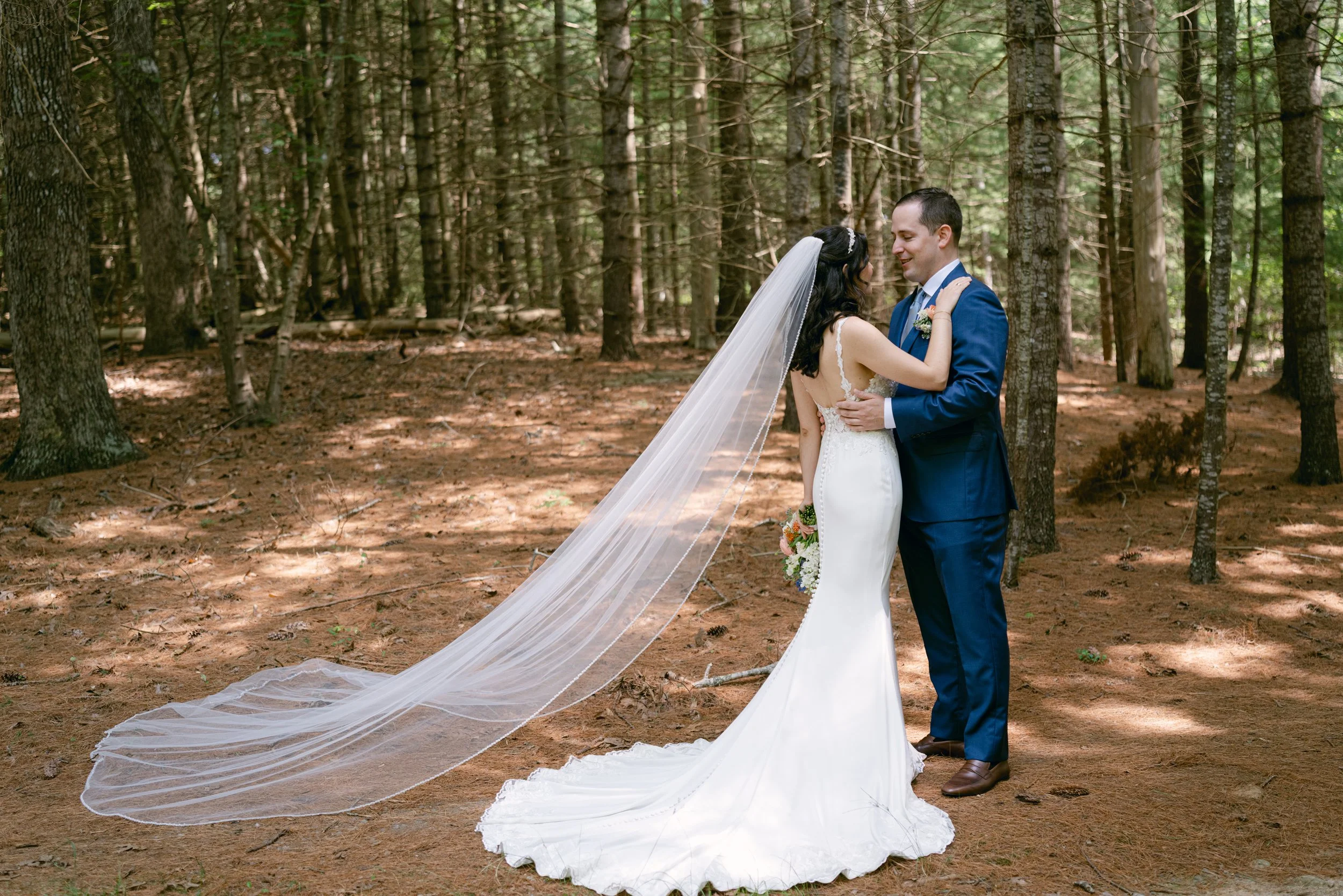 A bride and groom share a romantic moment in a forest, standing close and gazing into each other's eyes. The bride wears a white wedding gown with a long train and veil, while the groom wears a blue suit. Tall trees surround them, creating a natural,