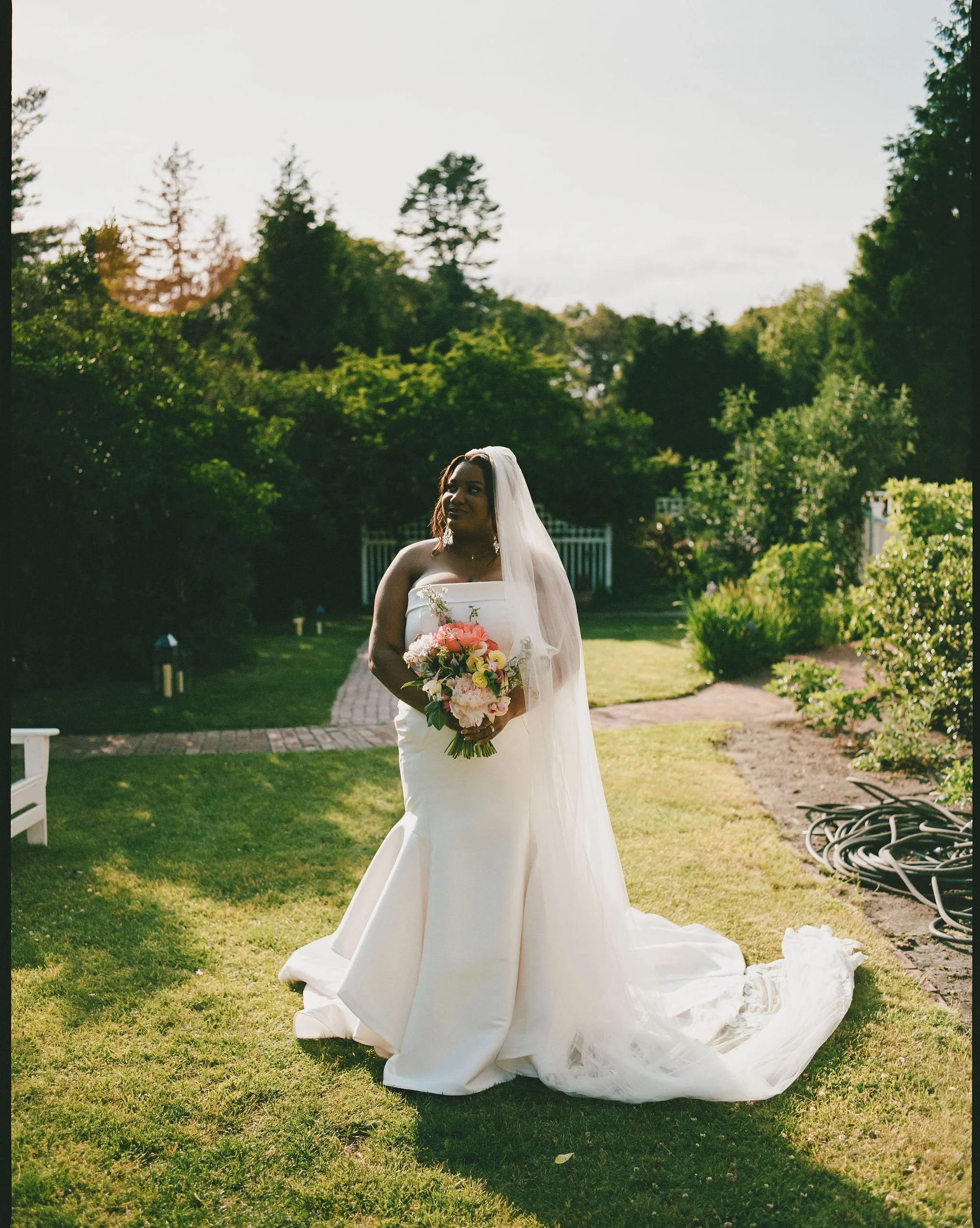 A bride in a white wedding gown and veil holding a bouquet of pink, white, and yellow flowers stands outdoors at Lambert's Cove Inn on Martha's Vineyard on a lush green lawn with trees in the background, and a white fence. The sky is partly cloudy.