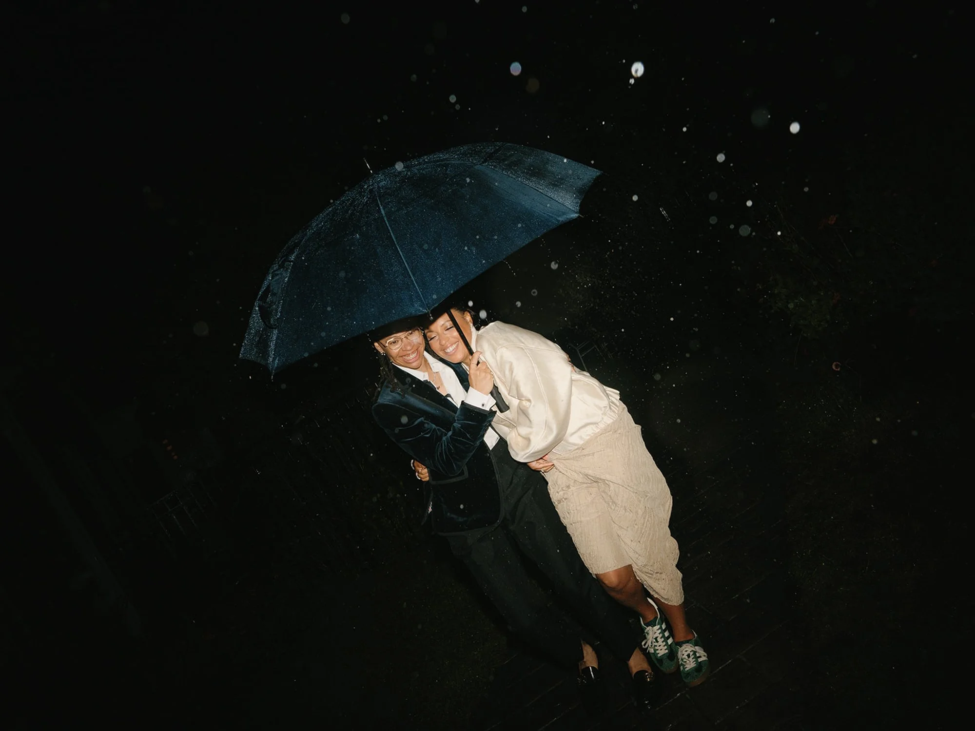 Two young women smiling and hugging under a blue umbrella in the rain at night.