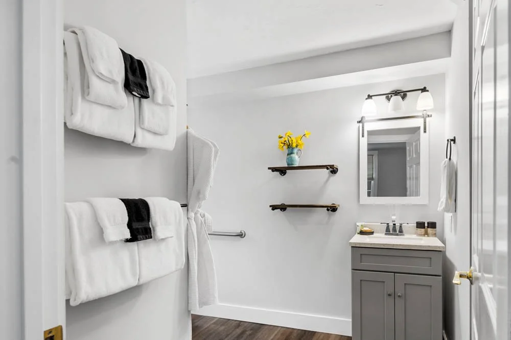 Bathroom with white towels on wall-mounted racks, a small gray vanity with a mirror, shelves with yellow flowers, and a towel hanging on a hook.