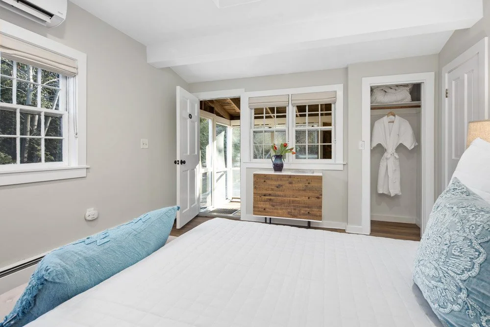 A bedroom with light gray walls, a window with white trim, a partially open door leading outside, a wooden wardrobe with a white robe hanging inside, a decorative vase with flowers on a wooden dresser, and a bed with white linens and blue decorative 
