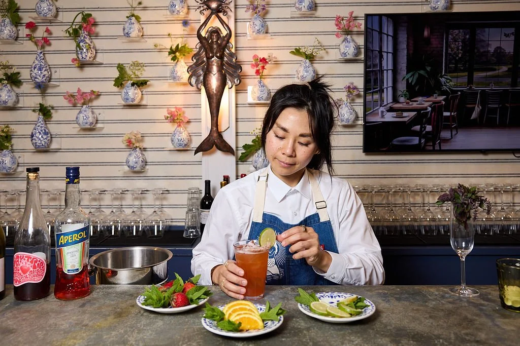 A woman preparing a cocktail at Mermaid Bar at Lambert's Cove Inn on Martha's Vineyard with blue and white vases and floral decorations on the wall behind her.