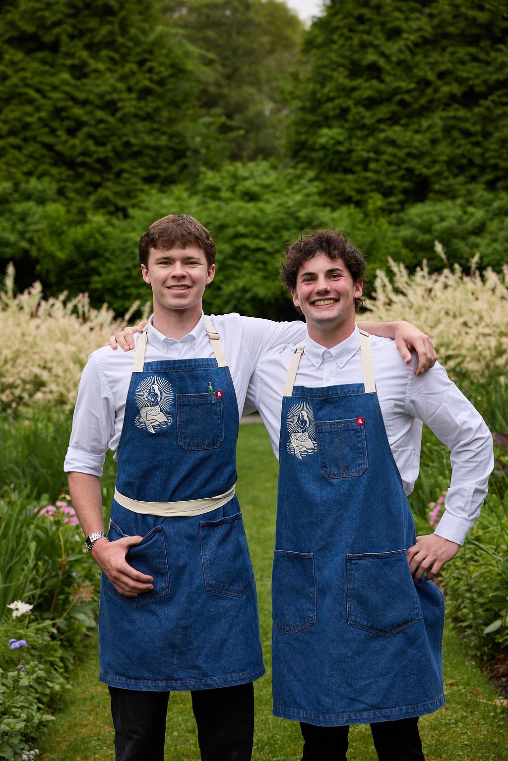 Two young men smiling and standing together in a garden, wearing white shirts and denim aprons with a religious logo, with lush trees and flowering plants in the background at Restaurant Foster at Lambert's Cove Inn on Martha's Vineyard.