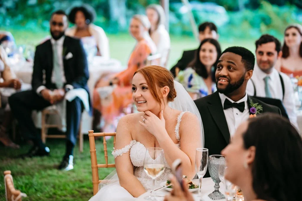 Bride and groom at their wedding reception at Lambert's Cove Inn on Martha's Vineyard, smiling and listening to speeches, surrounded by guests in formal attire outdoors.