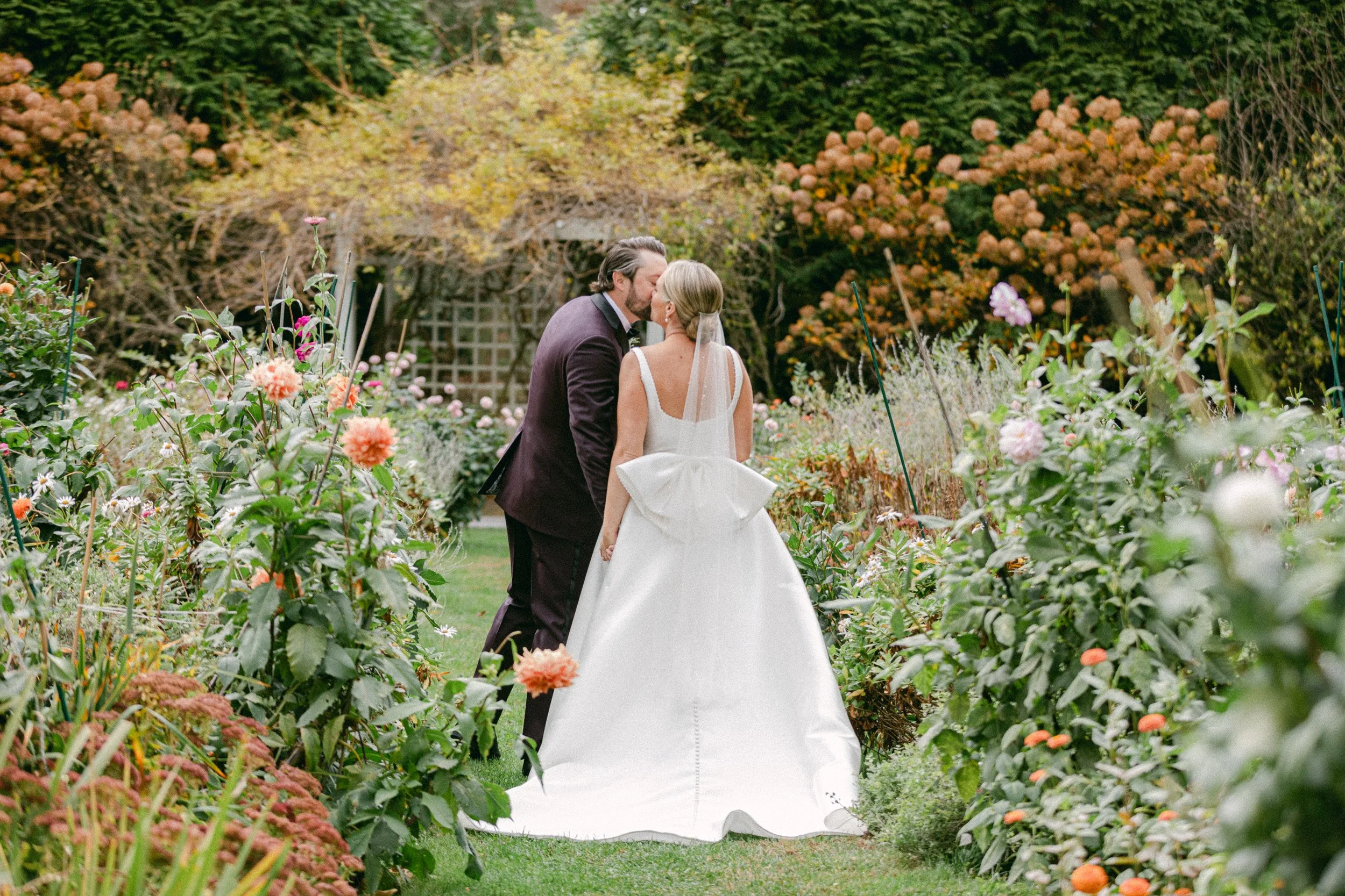 A bride and groom sharing a kiss in a garden surrounded by colorful flowers and greenery.