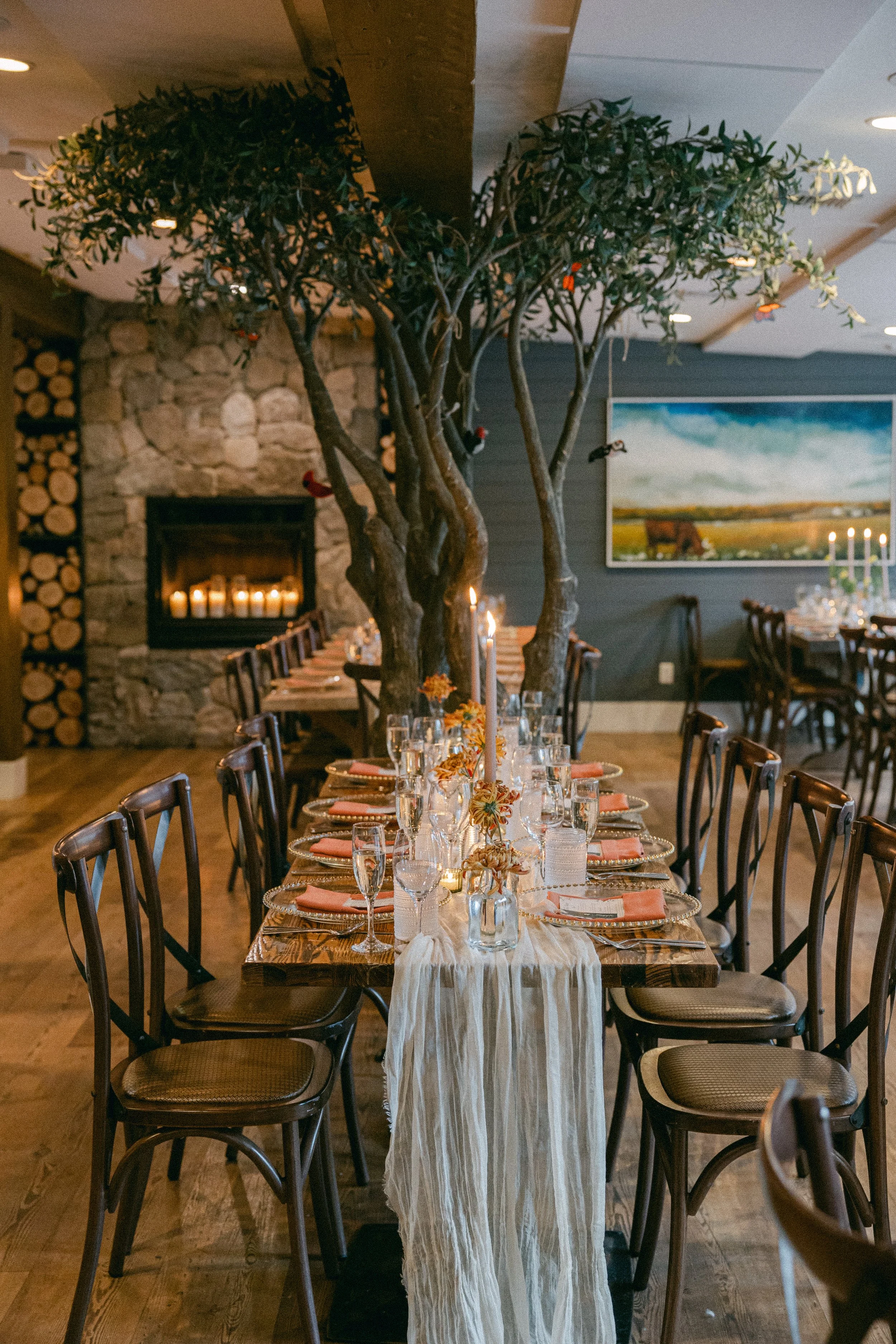 A long dining table set for a celebration, decorated with a white tablecloth, floral arrangements, and candles. A large indoor tree with multiple trunks stands in the center of the table, and a stone fireplace with candles burns in the background. Th