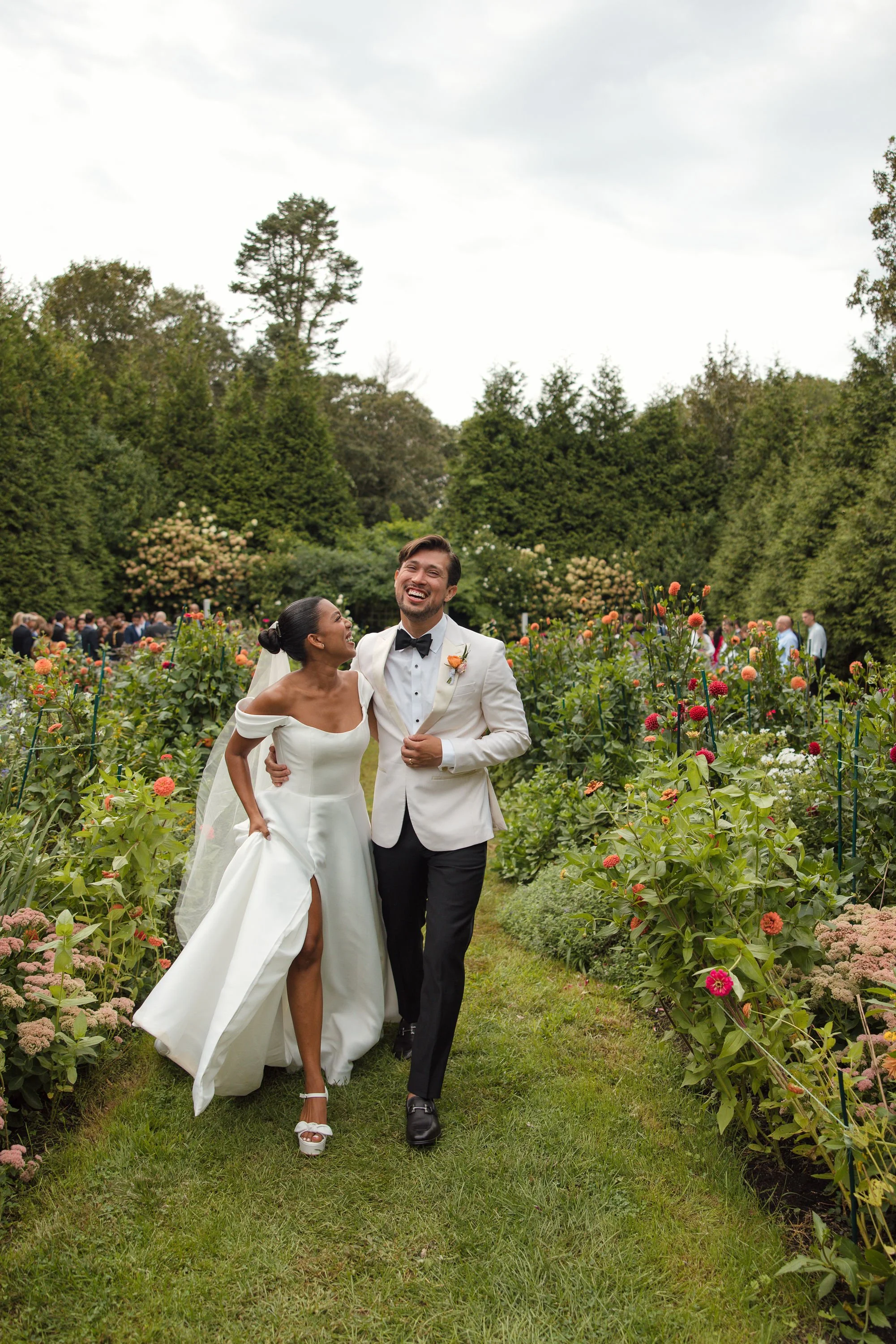 A newlywed couple walking through a flower garden, smiling and laughing, with wedding guests in the background during a wedding at Lambert's Cove Inn on Martha's Vineyard.