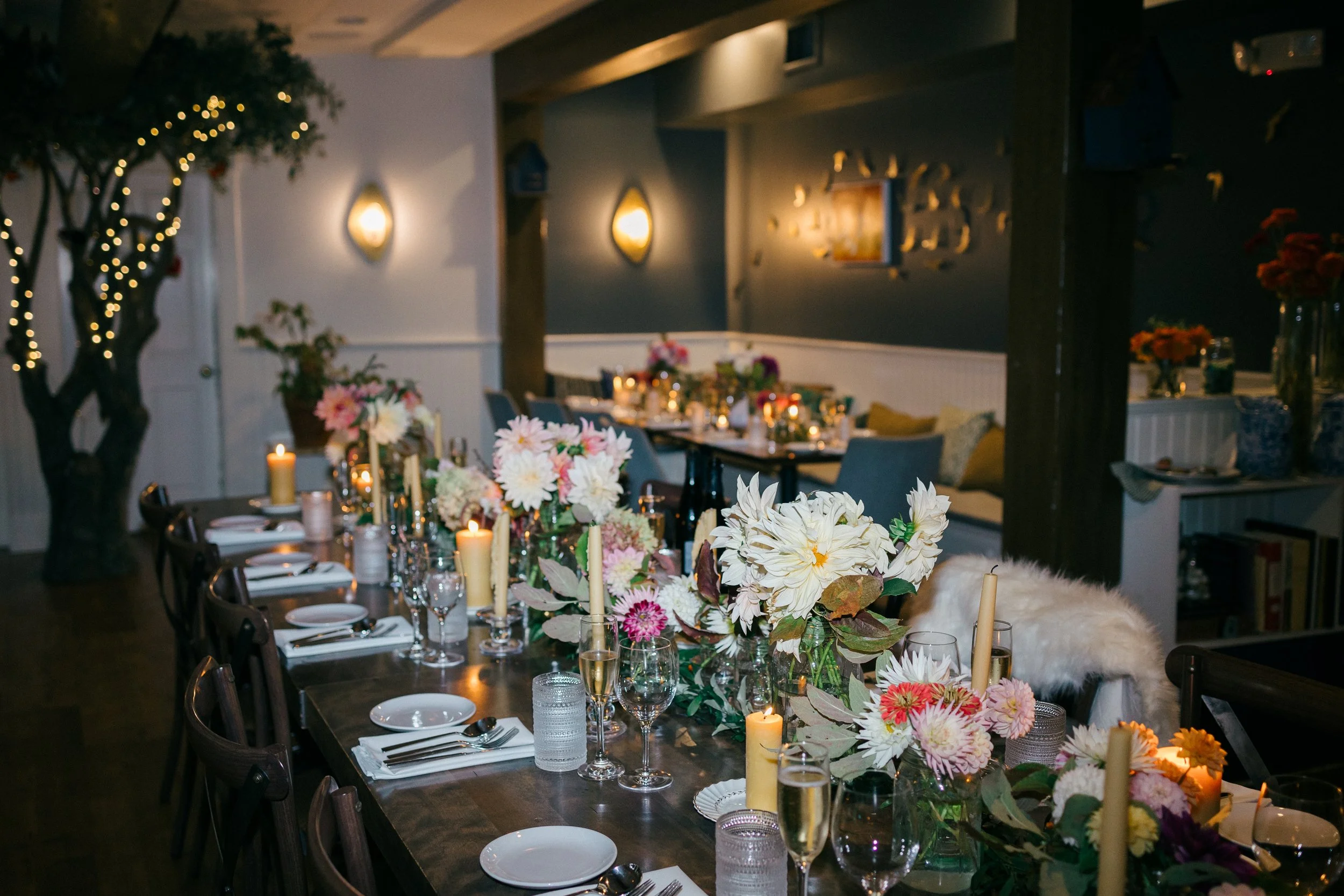 Indoor dining room decorated with flowers and candles on a long table, with wall-mounted lights and a wall art piece at Lambert's Cove Inn on Martha's Vineyard.