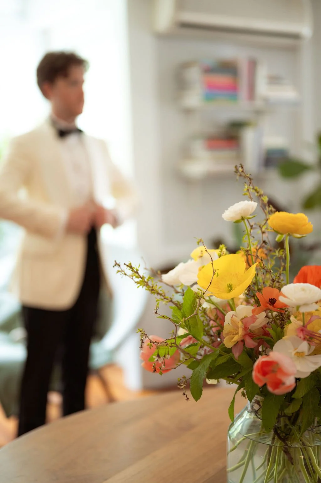 A bouquet of colorful flowers in a glass vase on a wooden table in a room with a blurred man in a tuxedo in the background at Lambert's Cove Inn on Martha's Vineyard.