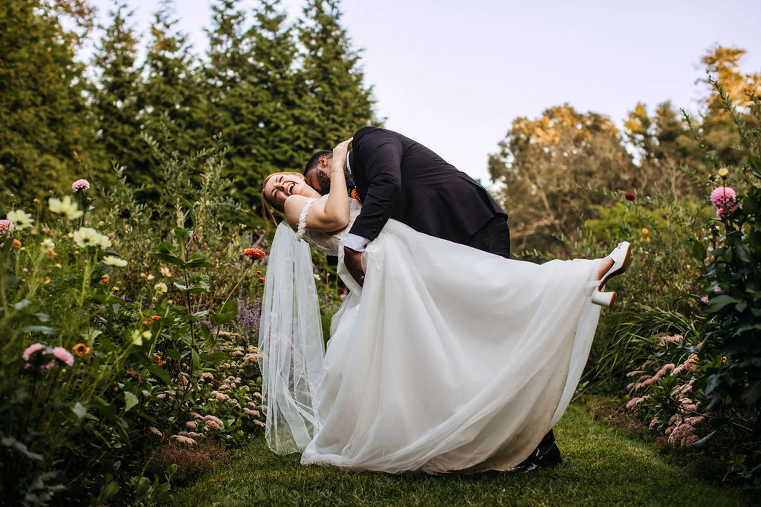 A newlywed couple is dancing and laughing in a garden with flowers and trees during their wedding celebration.