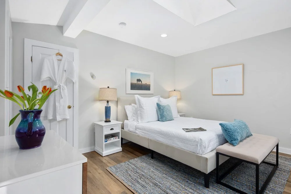 A bedroom with white walls, a bed with white bedding and blue pillows, a white nightstand with a blue lamp, framed artwork on the wall, a bench at the foot of the bed, a dresser with a blue flower vase, and a white robe hanging on the door.