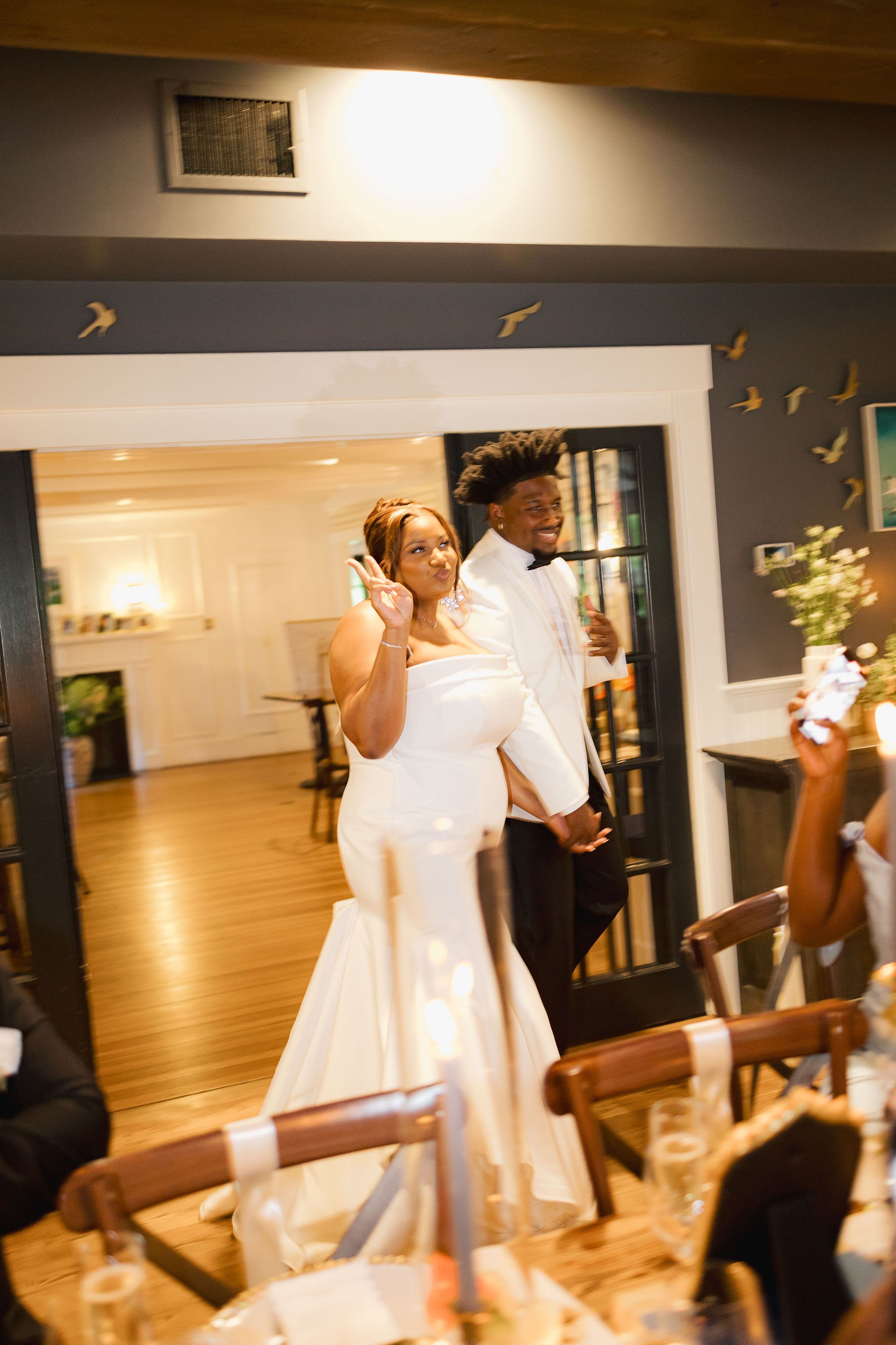 A woman in a white dress and a man in a white tuxedo pose for a photo, making peace signs at a celebration in a decorated room at Lambert's Cove Inn on Martha's Vineyard.