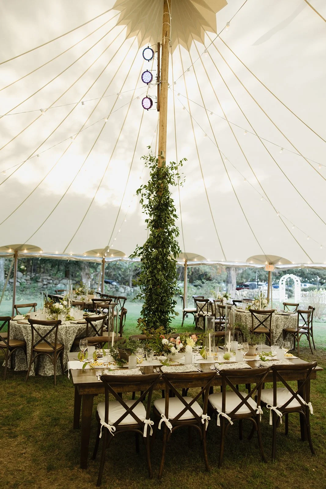 Indoor wedding reception at Lambert's Cove Inn on Martha's Vineyard setup under a large tent with decorated tables, chairs with bows, and a central pole covered in greenery and flowers, with string lights hanging from the ceiling.