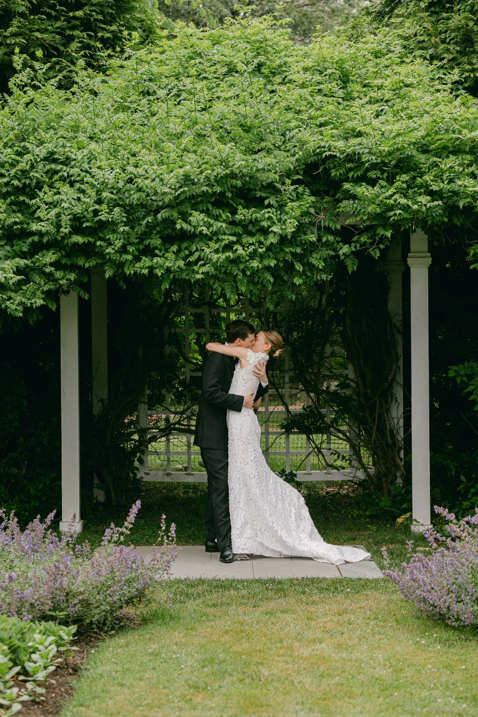 A bride and groom kissing outdoors under a large leafy green tree and arbor with purple flower border in a garden during a wedding at Lambert's Cove Inn on Martha's Vineyard.