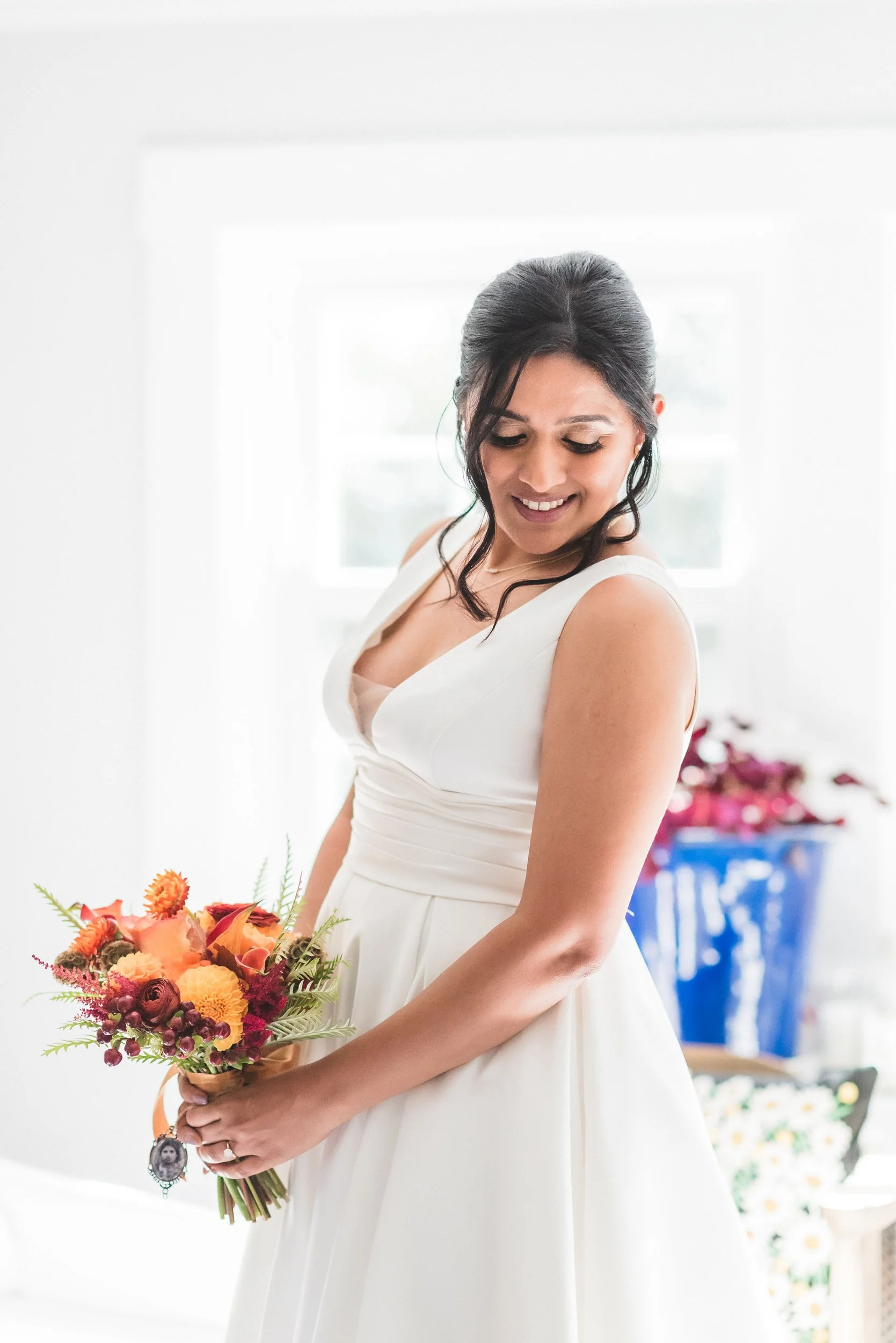 A woman in a white dress holding a colorful bouquet of flowers, smiling and looking down, in a bright room with a window in the background at Lambert's Cove Inn on Martha's Vineyard.