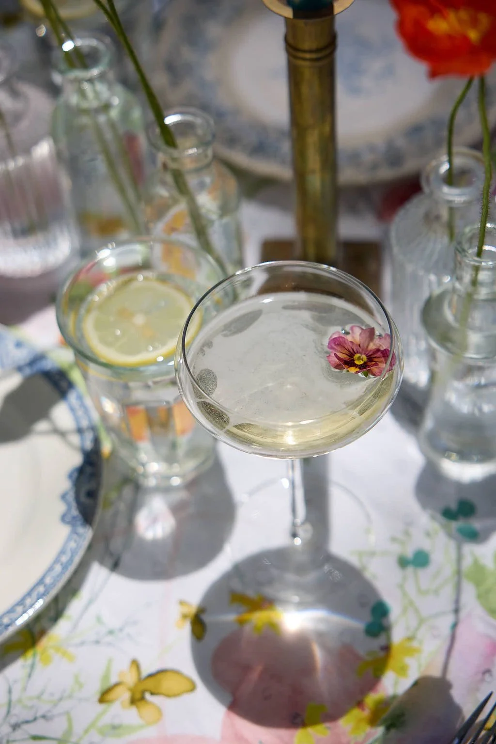 A cocktail glass with a flower floating on top, surrounded by bottles with lemon slices and flowers, on a table with a floral tableclothat Mermaid Bar at Lambert's Cove Inn on Martha's Vineyard.
