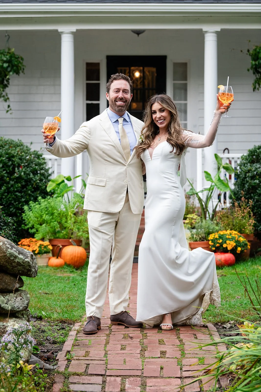 A newlywed couple in wedding attire celebrating outdoors in front of Lambert's Cove Inn on Martha's Vineyard, holding glasses of Aperol spritz.