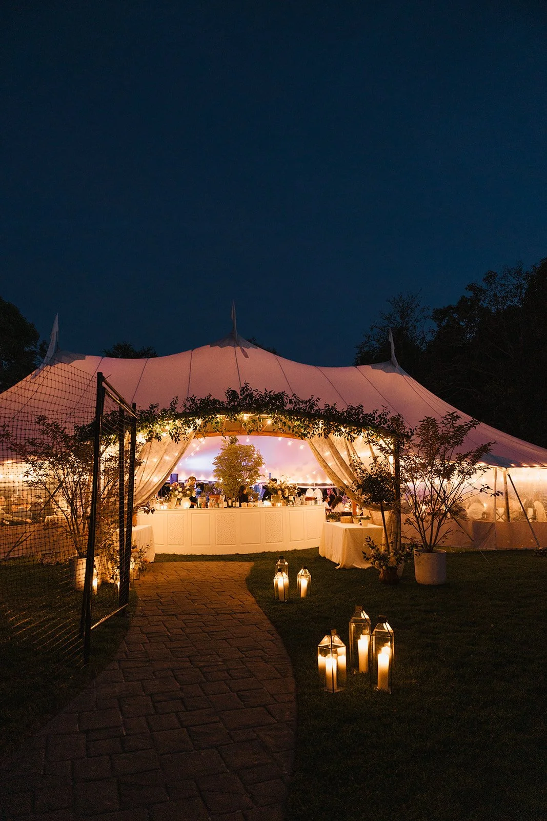 A large outdoor wedding tent illuminated with string lights at dusk, with lanterns along a brick pathway leading to the entrance, surrounded by trees and greenery at Lambert's Cove Inn on Martha's Vineyard.