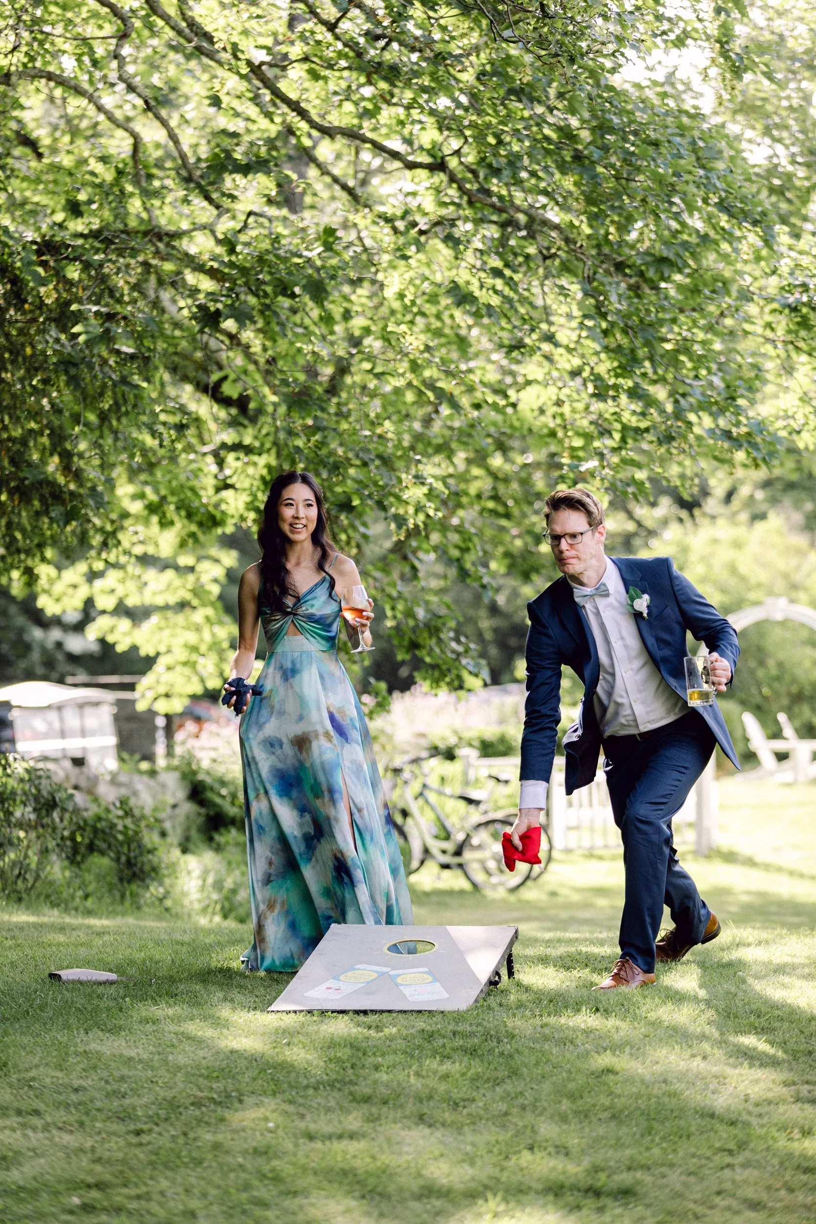 A woman in a colorful dress and a man in a tuxedo playing cornhole outdoors under a large tree. They both hold drinks and are engaged in the game, with a cornhole board and bean bags visible on the grass.