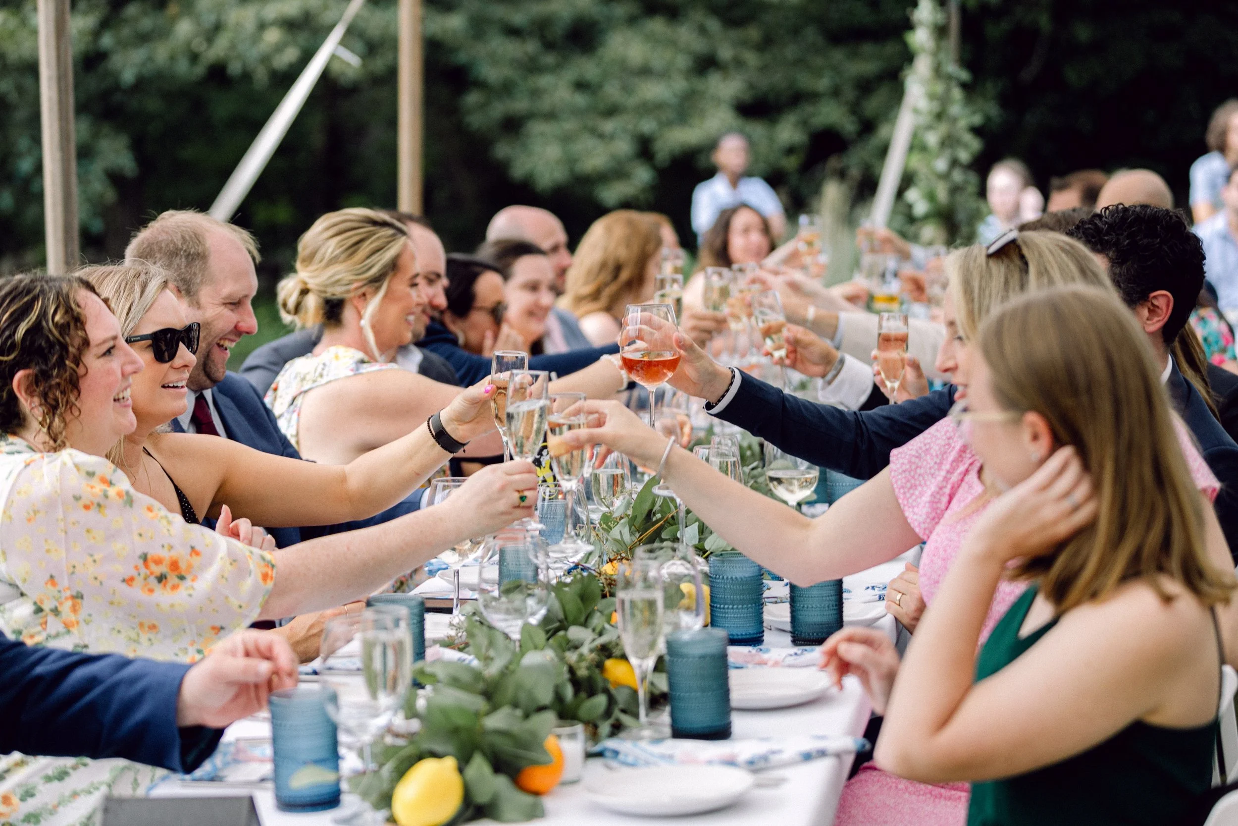 Wedding guests sitting at a long outdoor table raising glasses in a toast during a reception at Lambert's Cove Inn on Martha's Vineyard, with greenery in the background.