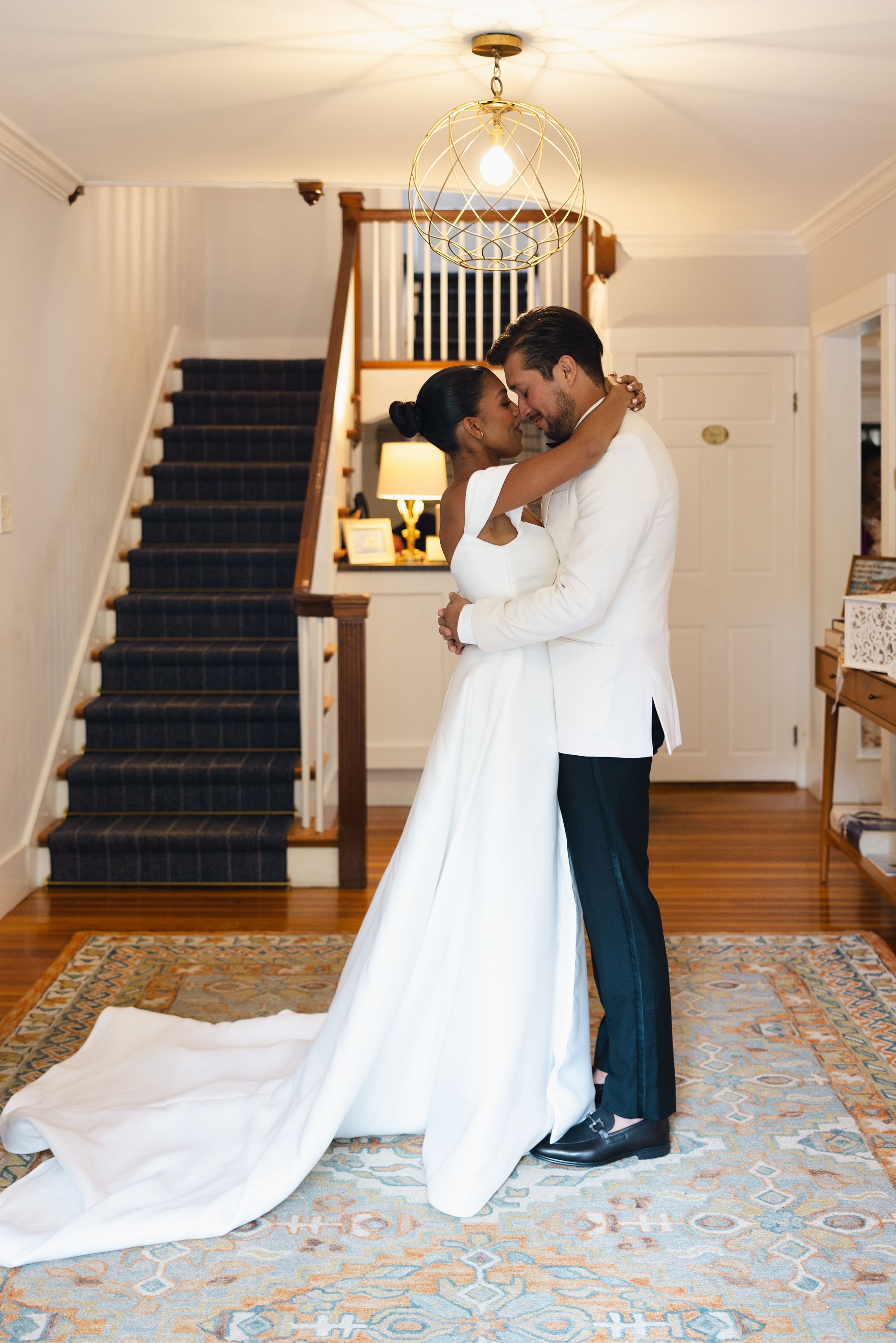 A bride and groom are standing close together indoors, embracing with their foreheads touching, during their wedding. The bride is wearing a white dress with off-the-shoulder sleeves, and the groom is in a white tuxedo jacket with dark pants. They ar