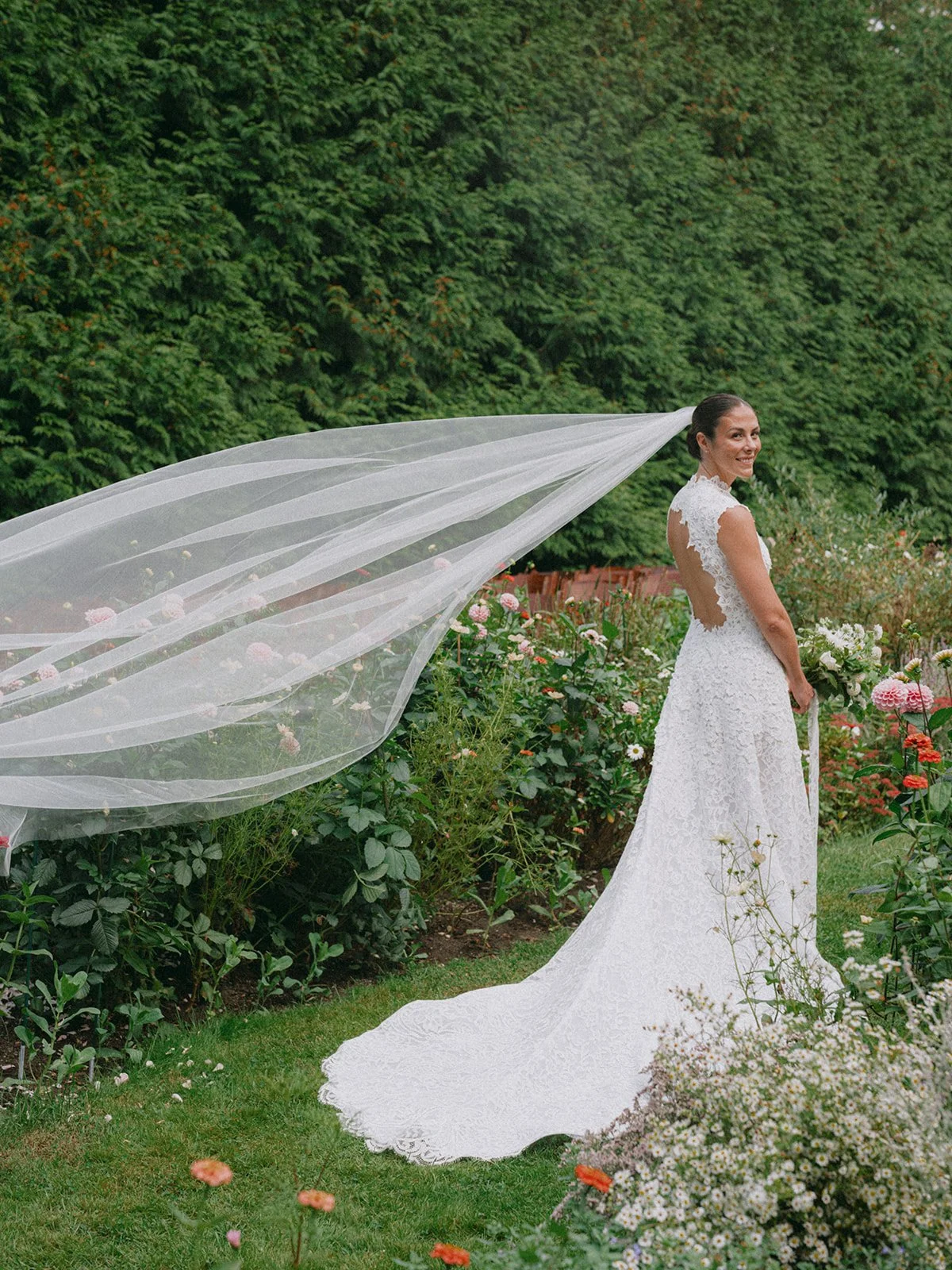 A bride in a white lace wedding dress standing outdoors in a garden with pink and white flowers. She is smiling, holding a bouquet, with a long flowing veil extending behind her. Tall green trees are in the background.