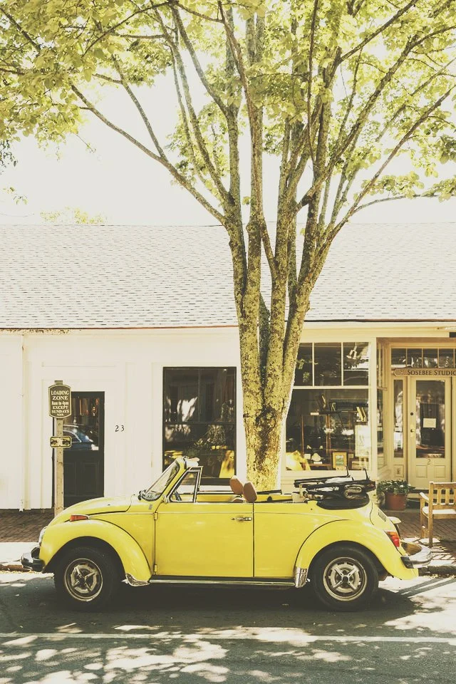 A yellow vintage convertible car parked on the street in front of a white building with large windows and a tree providing shade.