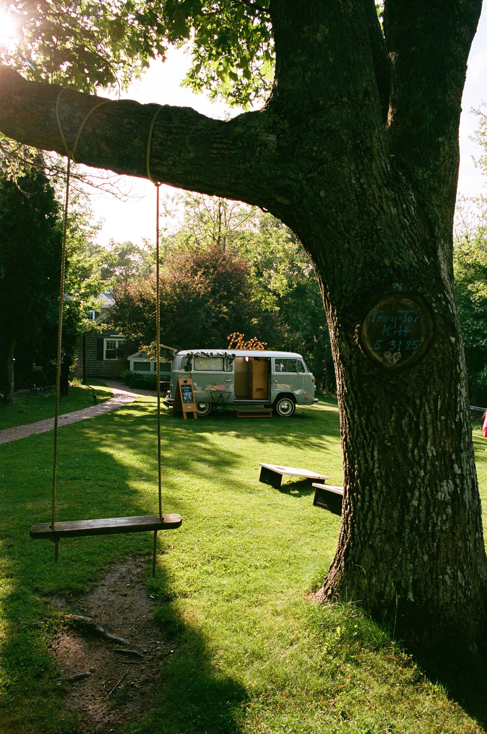 A large tree with a swing hanging from one of its branches, a dirt path leading to a park with lush green grass, a camper van in the background with a sign that says 'Photos', and a small chalkboard sign next to it.