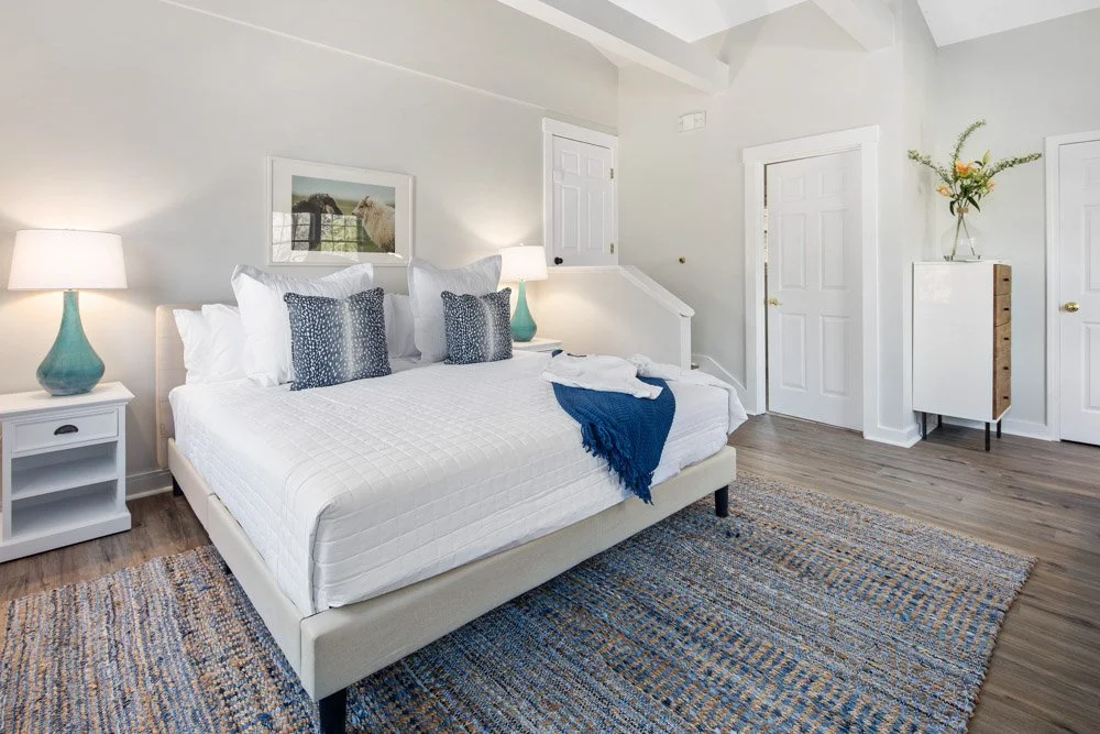 A bedroom with a white bed, four pillows, and a blue blanket. Two bedside lamps on white nightstands, a framed picture on the wall, wooden flooring, a multicolored rug, and a white cabinet with a flower vase.