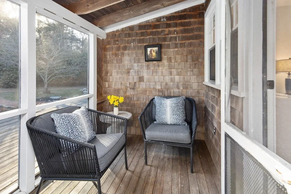 Enclosed porch with two black metal chairs with grey cushions and patterned pillows, a small side table with a vase of yellow flowers, and a framed picture on a shingled wall, overlooking a leafless tree outside.