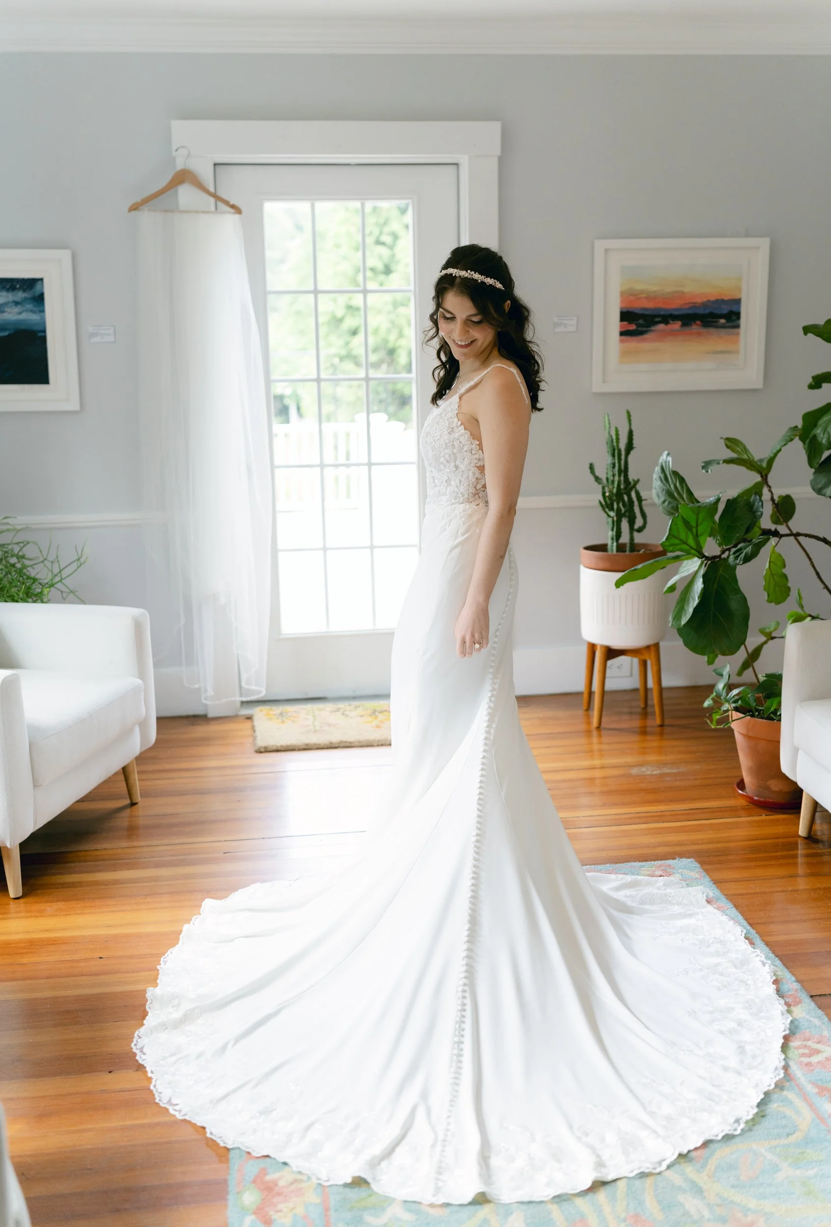 A woman in a wedding dress with a long train is standing in a brightly lit room, smiling gently. The room has a hardwood floor, white arm chairs, green plants, and framed artwork on the walls. There is a window with a view of greenery outside.