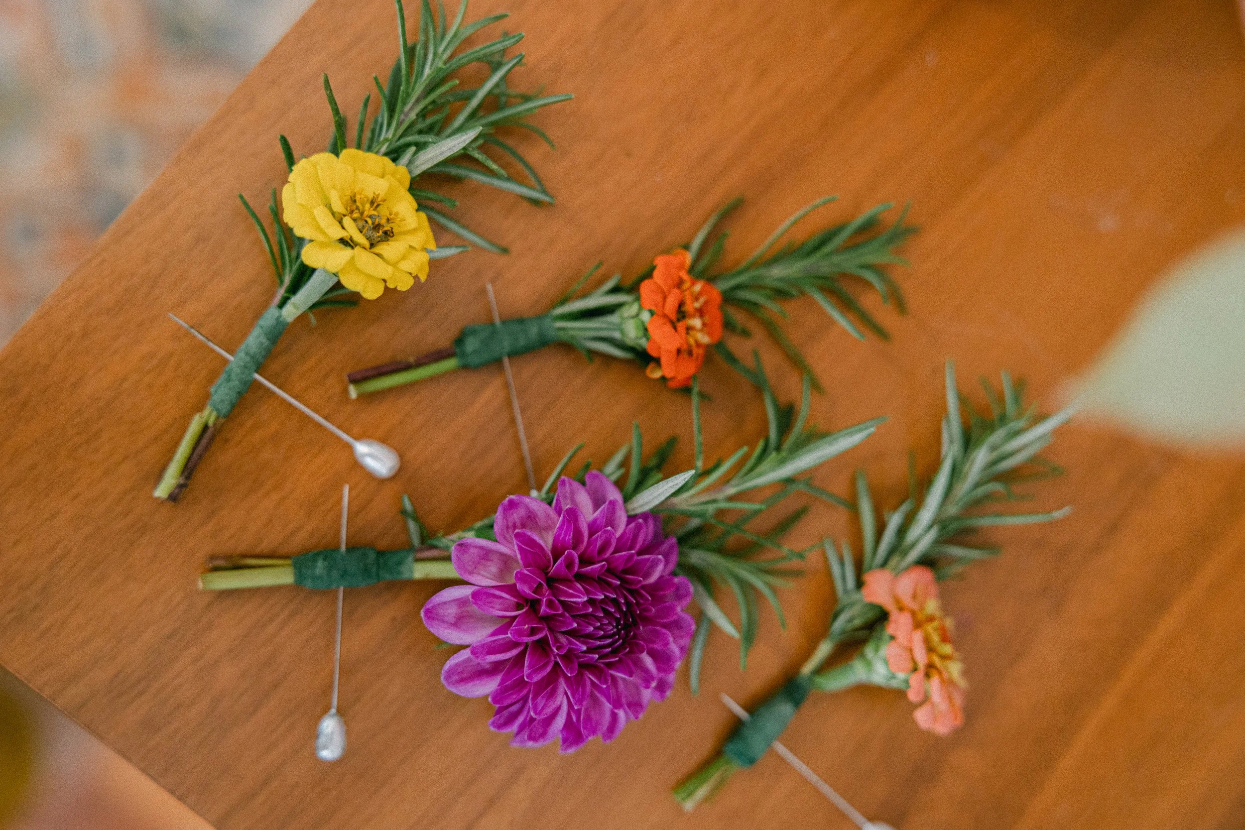 Colorful floral boutonnieres with yellow, orange, purple, and pink flowers, decorated with green leaves, lying on a wooden surface. The flowers were custom grown in the garden at Lambert's Cove Inn on Martha's Vineyard.