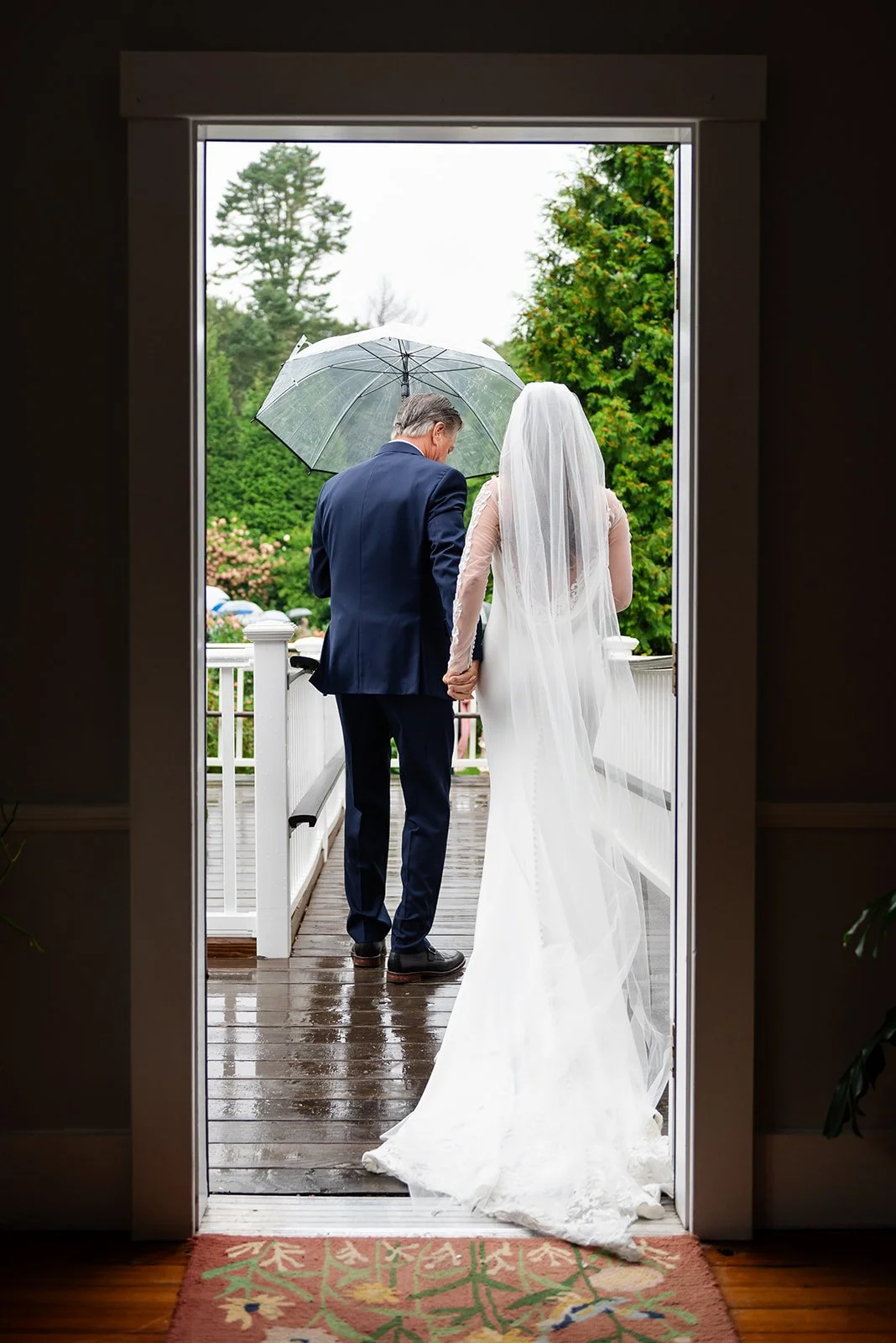 A bride and groom holding hands on a rainy day outside, viewed through a doorway, with the groom holding an umbrella at Lambert's Cove Inn on Martha's Vineyard.