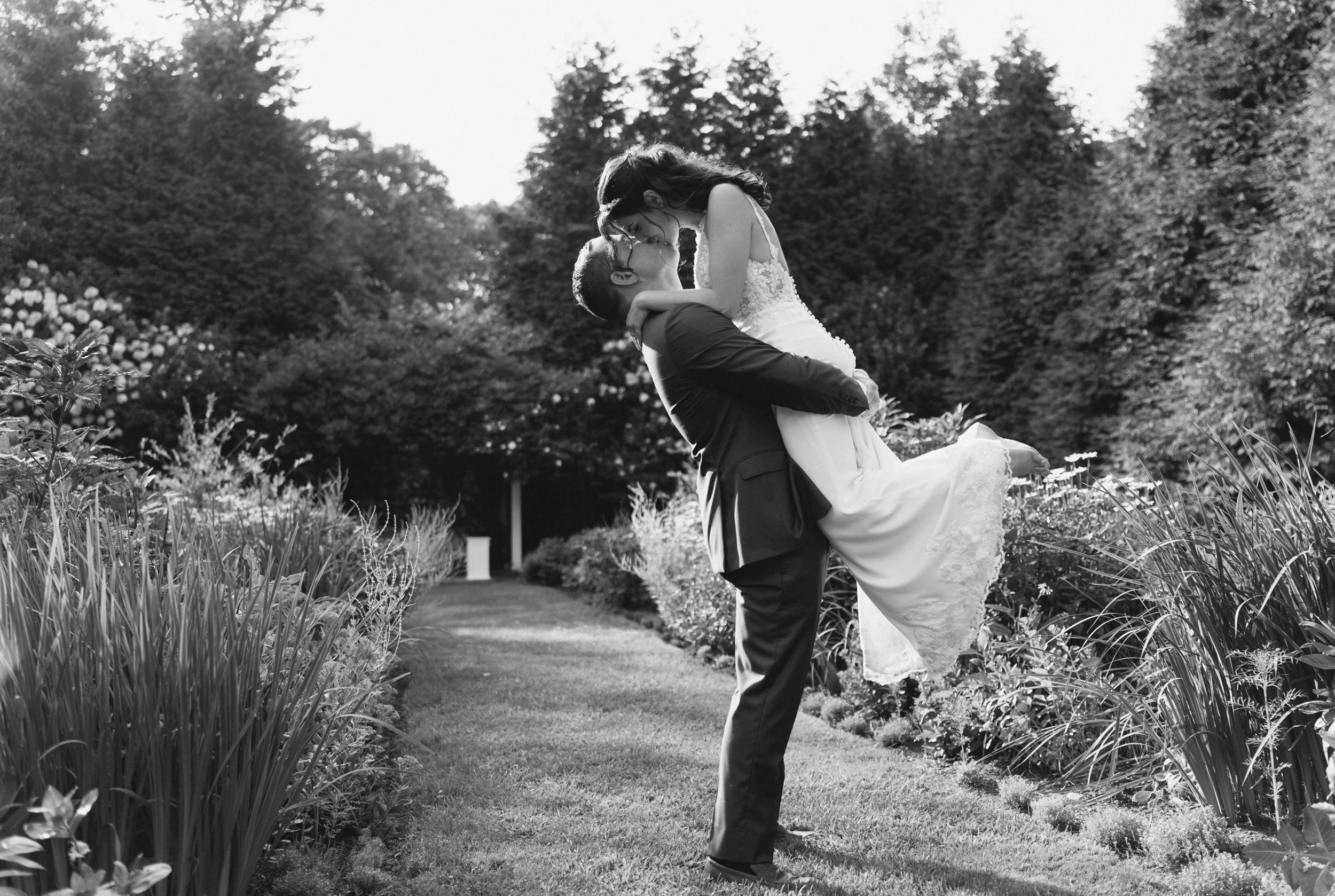 A black-and-white photo of a couple, with the man lifting and kissing the woman in a garden surrounded by trees and plants, with a pathway in the background.
