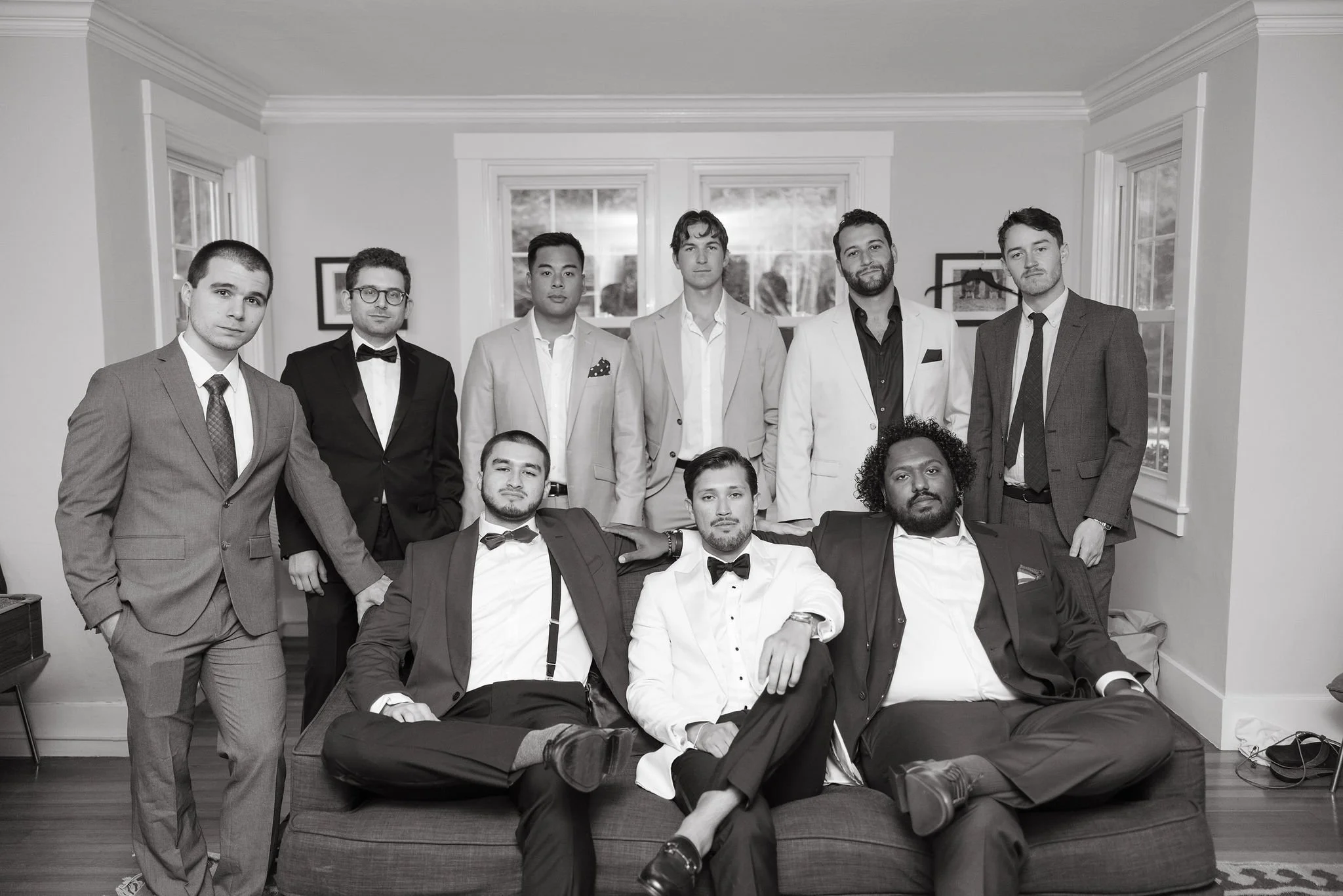 Group of nine men dressed in formal suits and tuxedos, some sitting and others standing, in a living room with large windows at Lambert's Cove Inn on Martha's Vineyard.