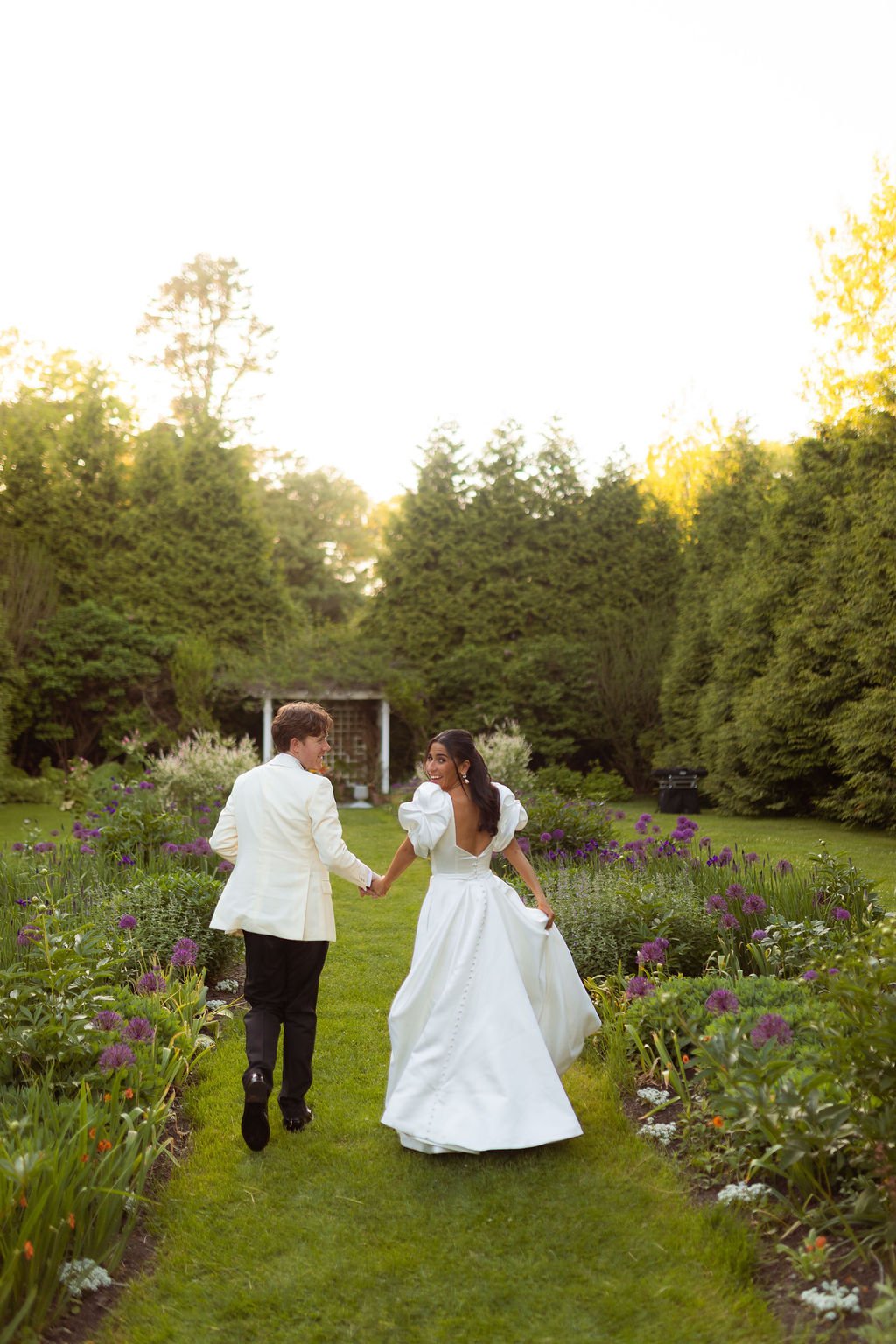 A newlywed couple walking hand in hand through the lush English garden at Lambert's Cove Inn on Martha's Vineyard, amid flowering plants, with the bride in a white wedding gown and the groom in a white suit, smiling at each other during sunset.