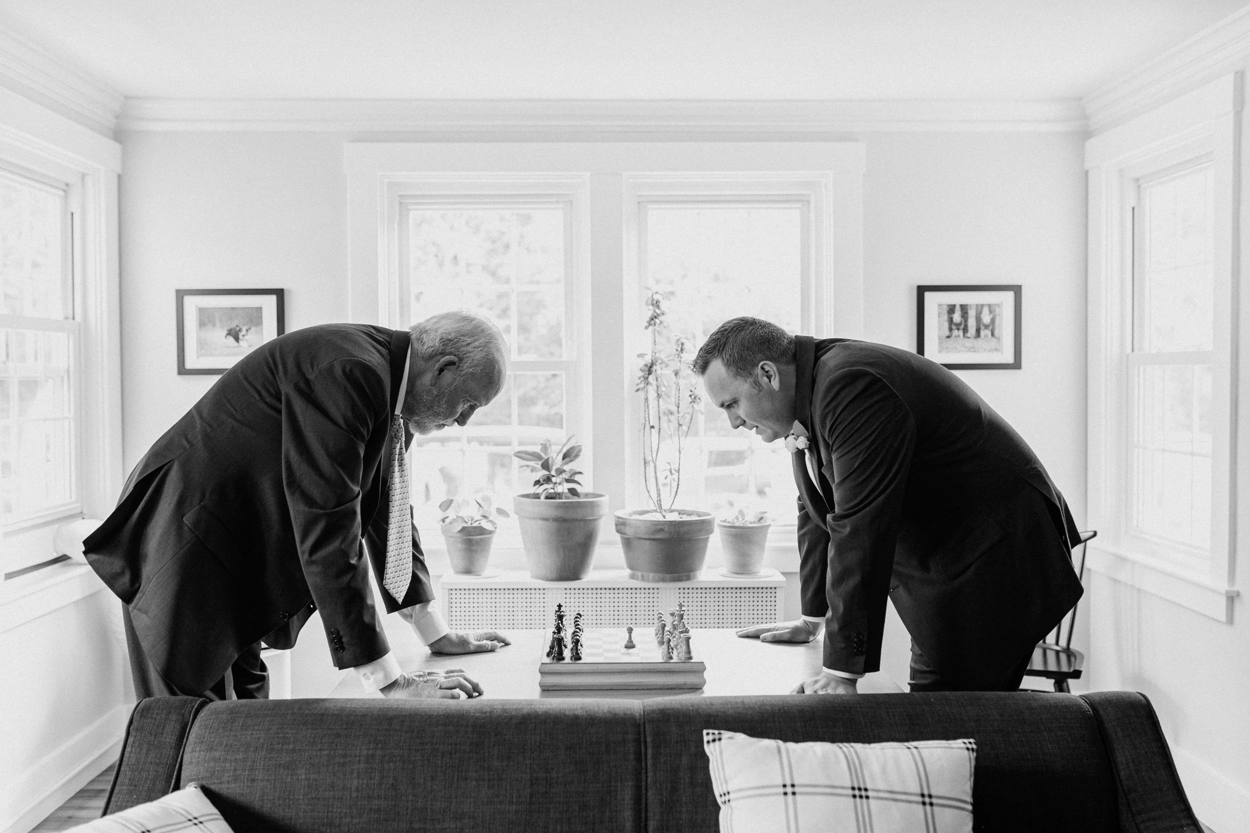 Two men dressed in suits playing chess in a bright room with large windows, framed pictures on the walls, and potted plants on a table before a wedding at Lambert's Cove Inn on Martha's Vineyard.
