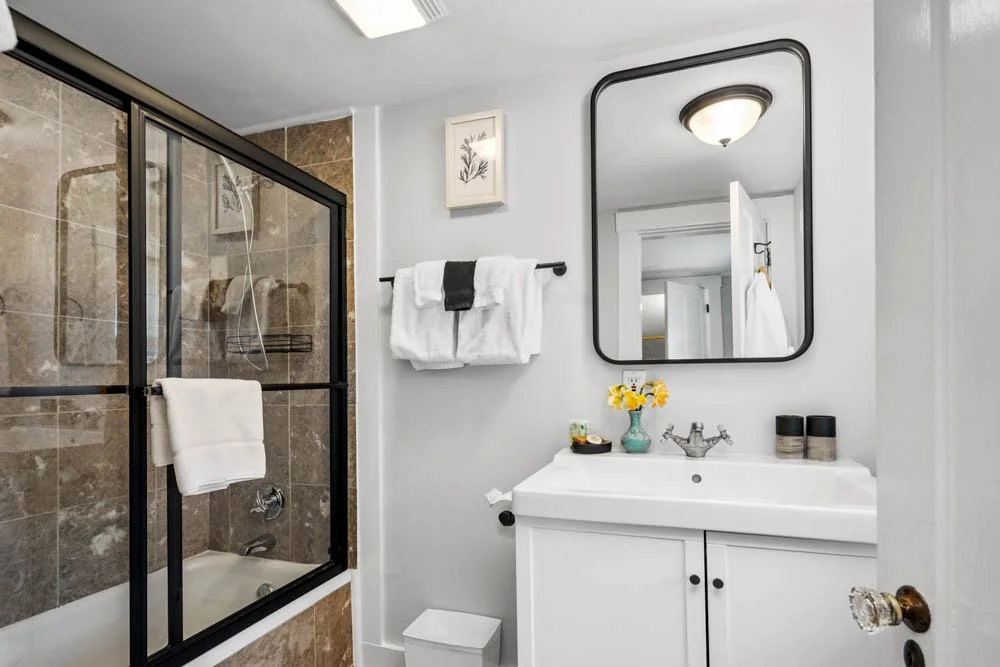 Bathroom at Lambert's Cove Inn with a glass-enclosed shower, a white sink with a cabinet, a mirror, a towel rack with towels, and a small decorative vase with yellow flowers.