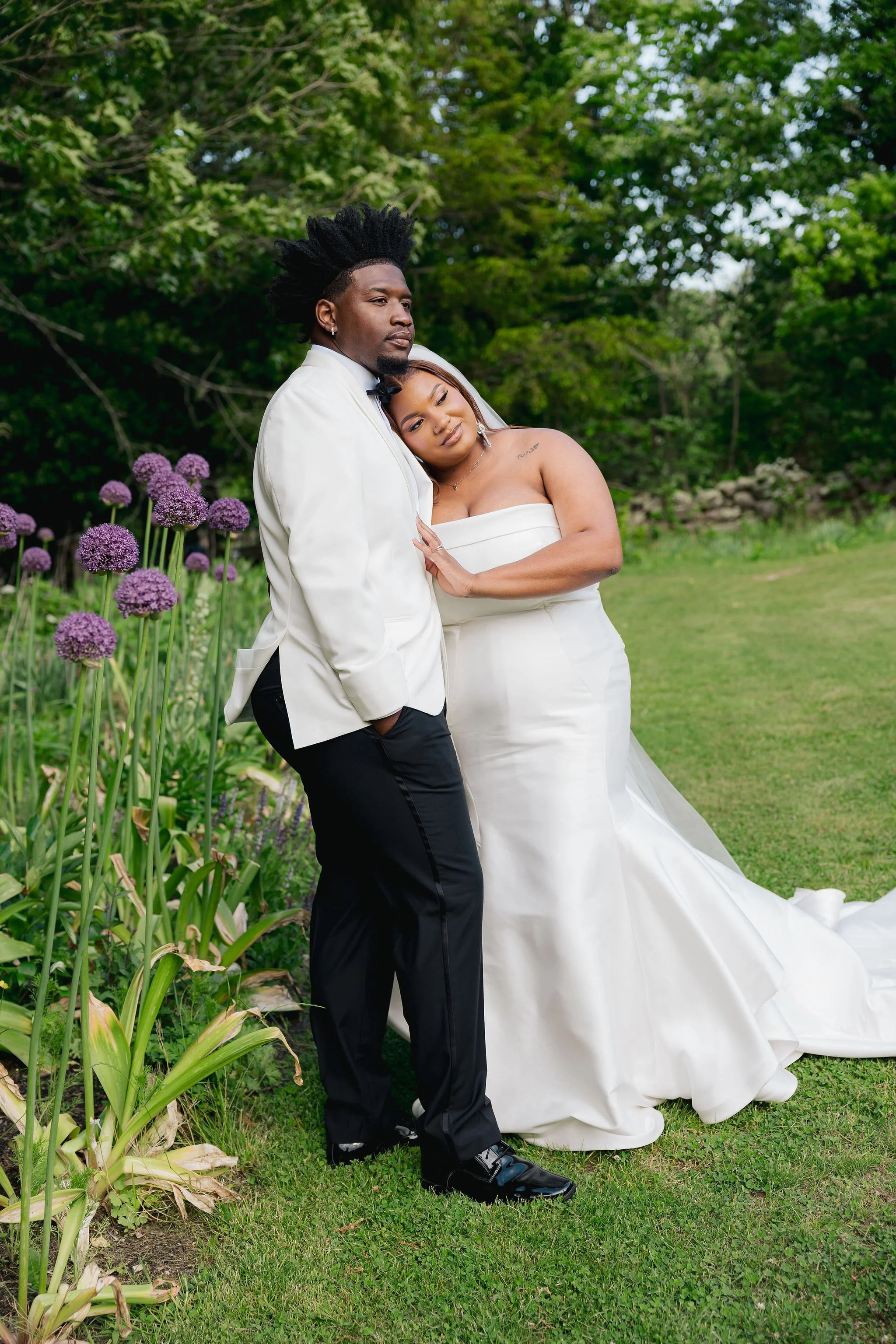 A bride and groom embrace outdoors in a garden with purple flowers and green trees, both wearing wedding attire. The woman leans her head on the man's chest, smiling gently, while the man stands tall in a white tuxedo jacket with black pants.