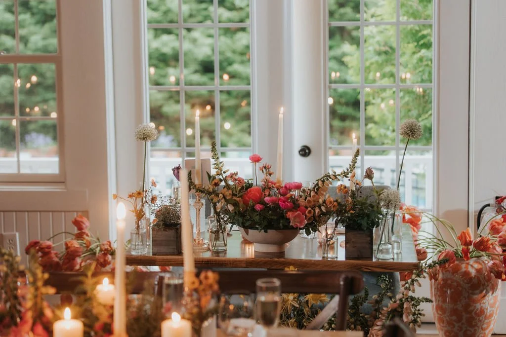 A decorated dining table with floral arrangements, lit candles, and glassware, set near large windows overlooking a green outdoor space.