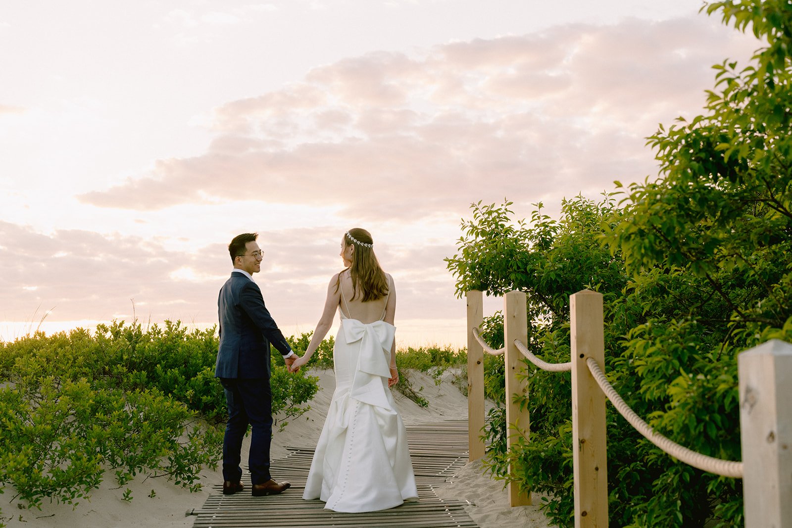 A bride and groom holding hands and walking on a beach pathway surrounded by greenery during sunset during a wedding at Lambert's Cove Inn on Martha's Vineyard.