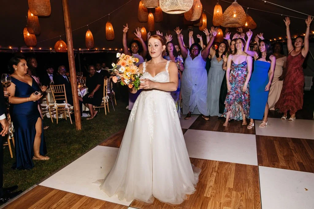 Bride in white wedding gown holding a bouquet of flowers in front of dancing guests during a wedding reception at Lambert's Cove Inn on Martha's Vineyard.