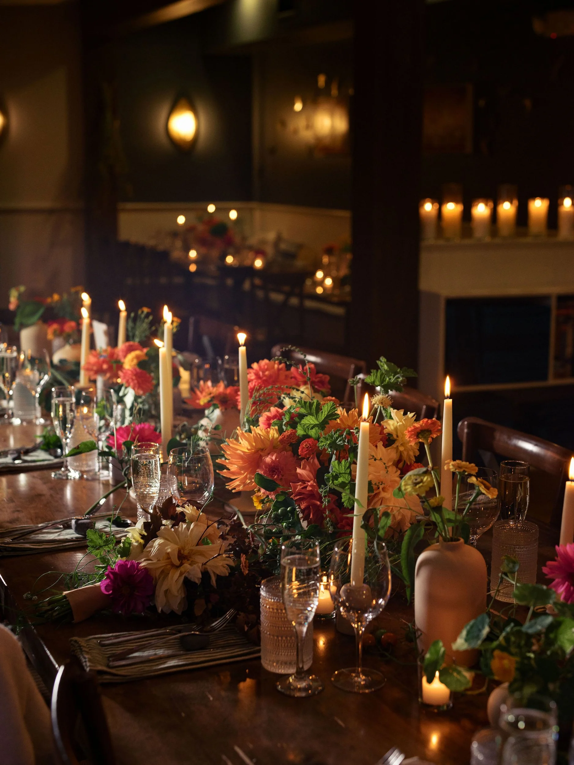 A dimly lit restaurant or dining room with a long table decorated with vibrant flower arrangements and tall white candles. The background features warm wall-mounted lights and a cozy, intimate atmosphere.
