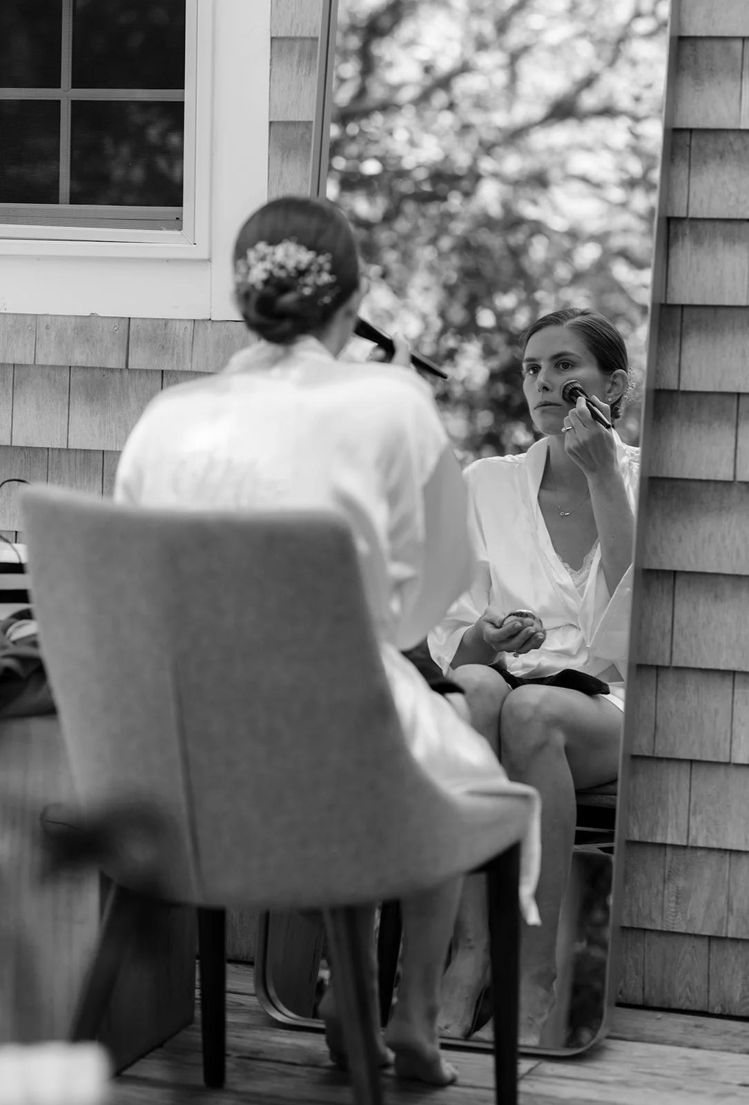 A bride applying makeup while looking into a mirror outside on a porch, with a woman sitting with her back to the camera in the foreground, prior to her wedding at Lambert's Cove Inn on Martha's Vineyard.