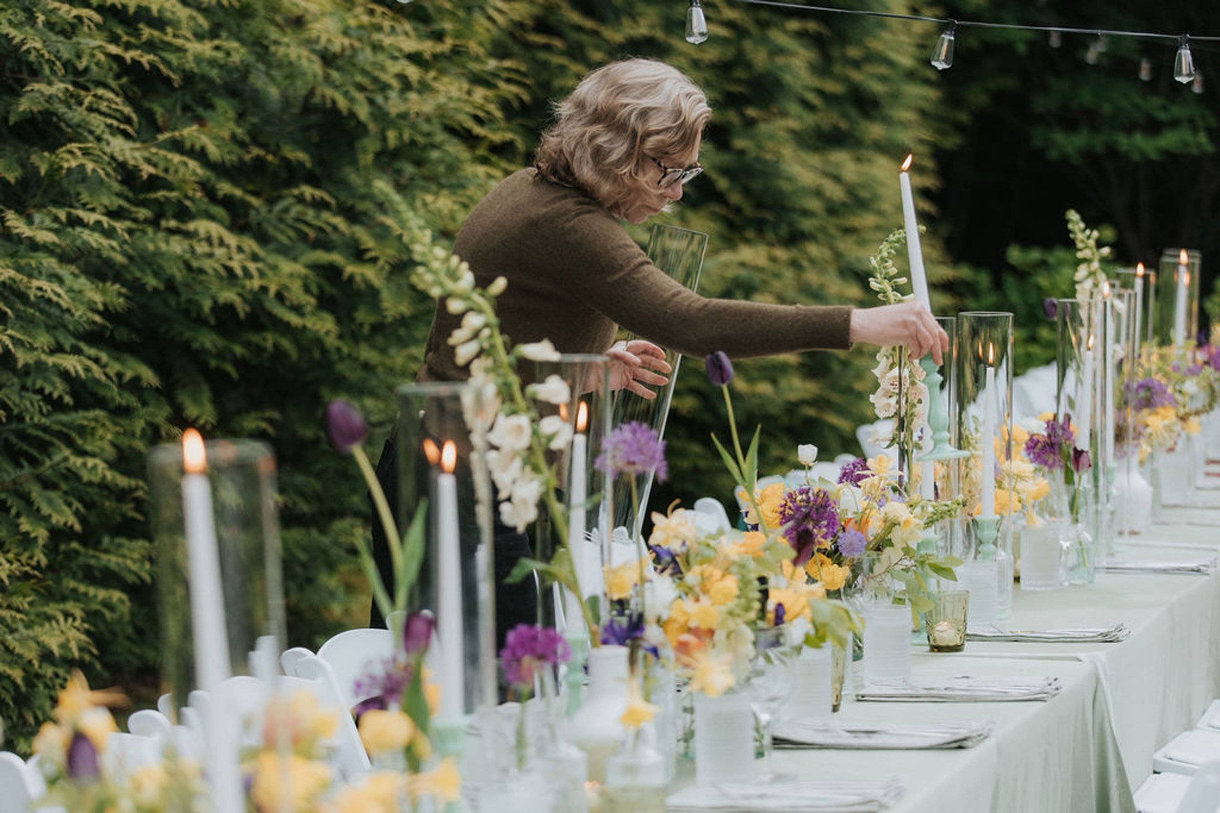 A woman, Bridget Sampson, the owner of at Lambert's Cove Inn on Martha's Vineyard, is decorating a table outdoors with flowers and candles, surrounded by greenery for an outdoor wedding reception in West Tisbury, MA.