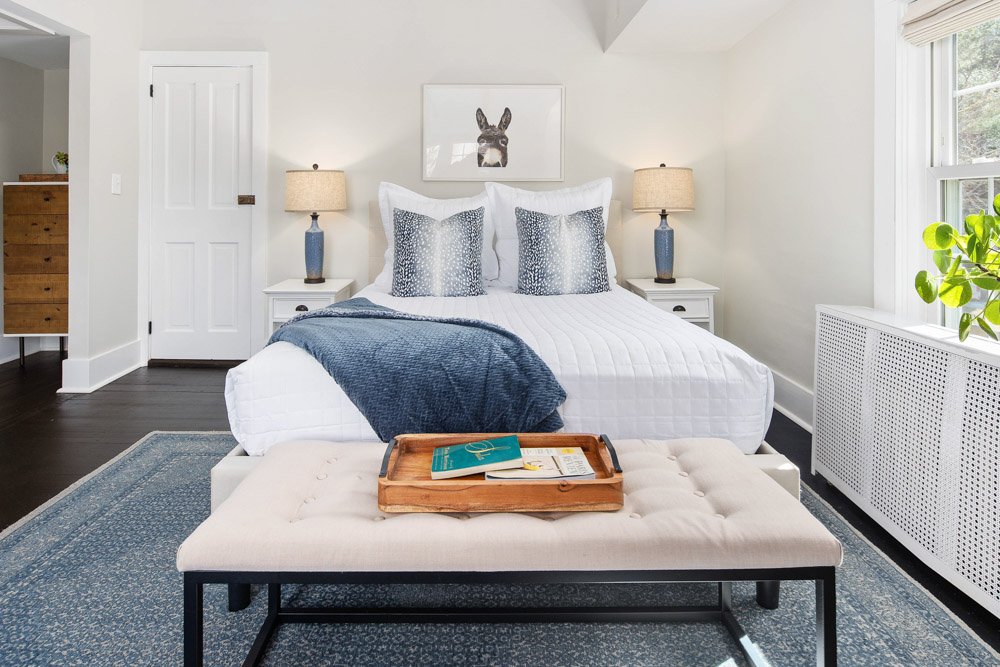 Guest room at Lambert's Cove Inn on Martha's Vineyard with a bed, white bedding, blue pillows, navy throw blanket, side tables with lamps, framed bunny picture, window with blinds, and a bench with a tray holding books and papers.
